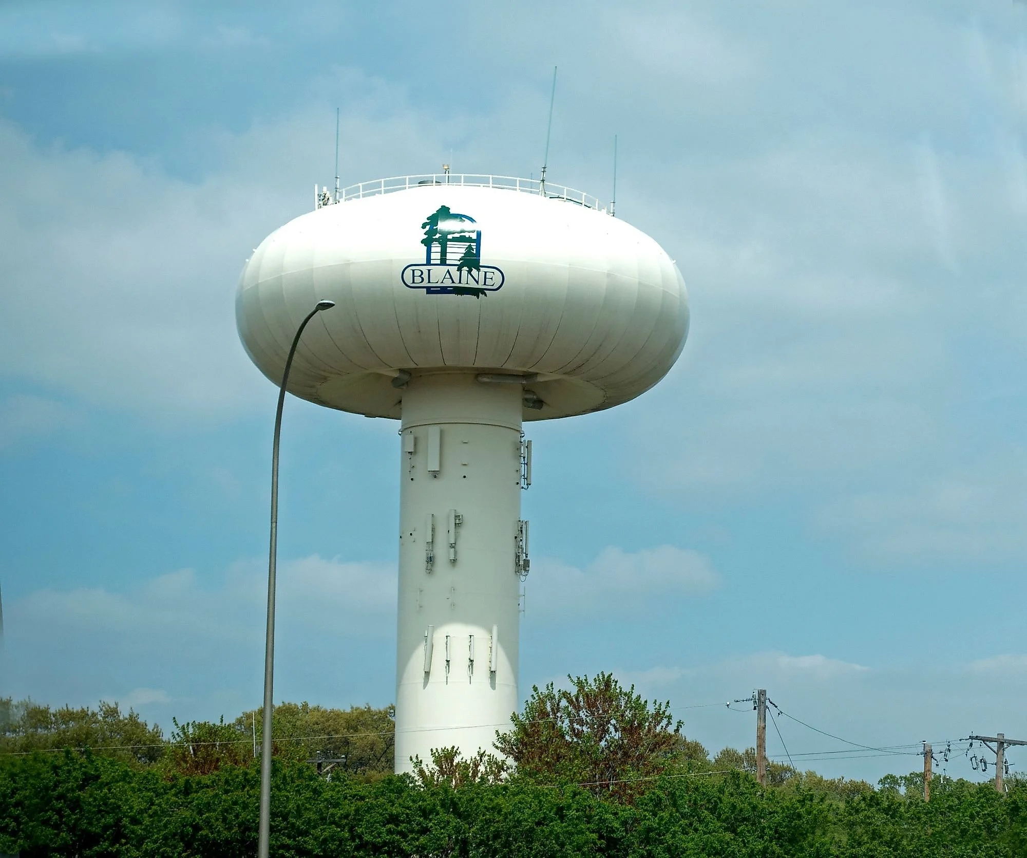 A tall, white water tower with a logo that says 'Blaine' and features a silhouette of a cowboy on a horse. There are trees, utility poles, and power lines in the foreground, and the sky is cloudy.