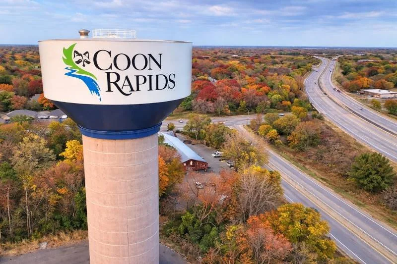 A water tower with the logo and name of Coon Rapids, featuring a green and black orca and a blue wave, with a background of trees showing fall foliage and a highway.