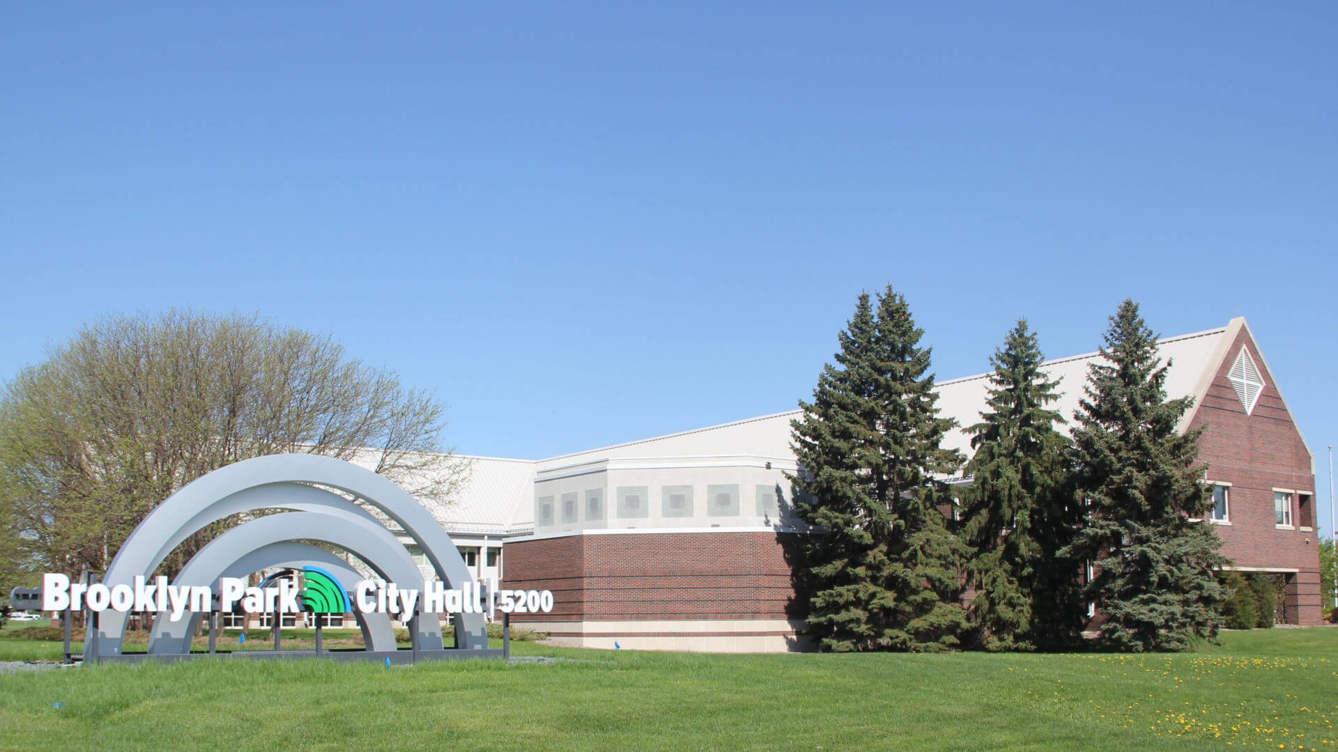 Sign for Brooklyn Park City Hall with trees and a building in the background under a clear blue sky.