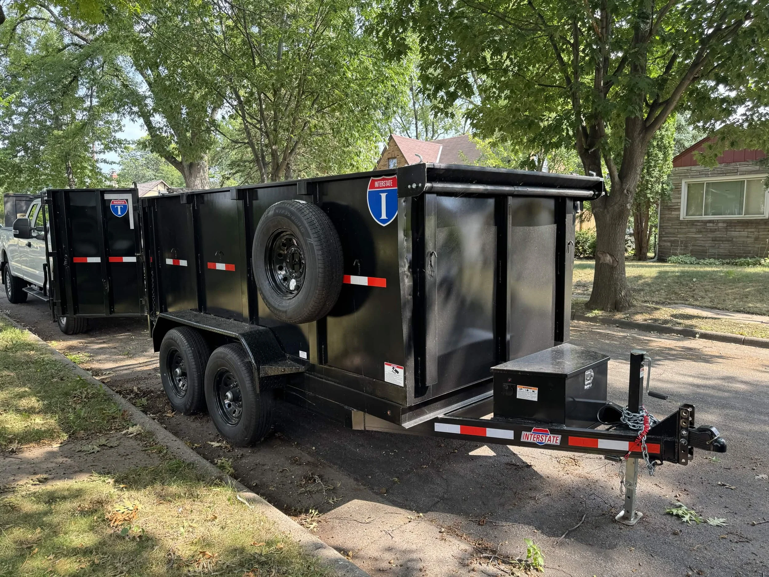 Black dump trailer attached to a pickup truck parked on a residential street with large trees and houses in the background.