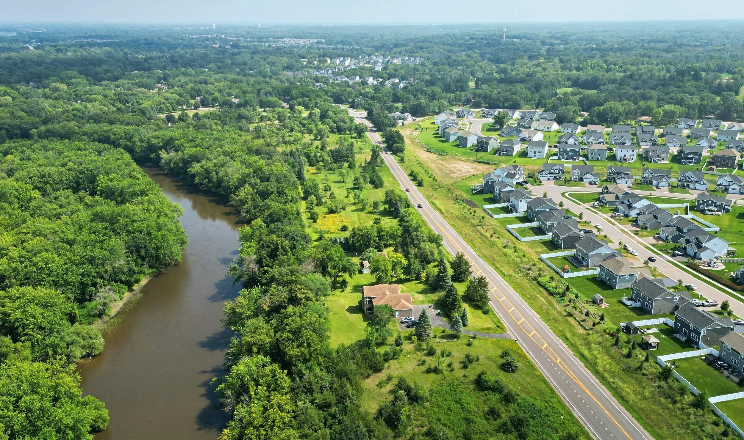 Aerial view of a residential neighborhood next to a river with dense green trees and houses along a road.