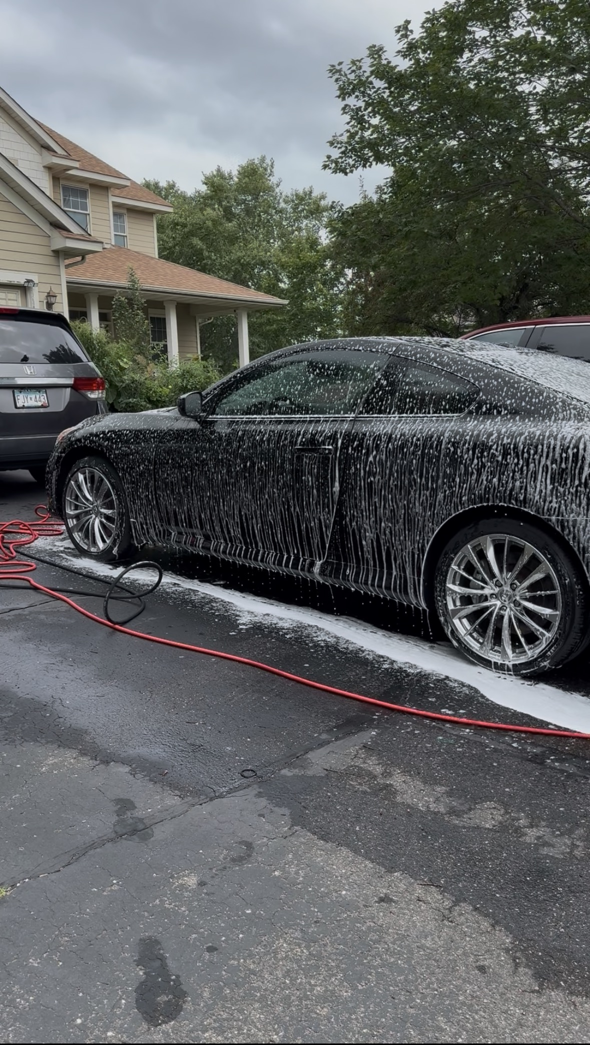 Black sports car being washed with soap and water on a driveway, with a garden hose connected to the car, and a house with trees and cloudy sky in the background.