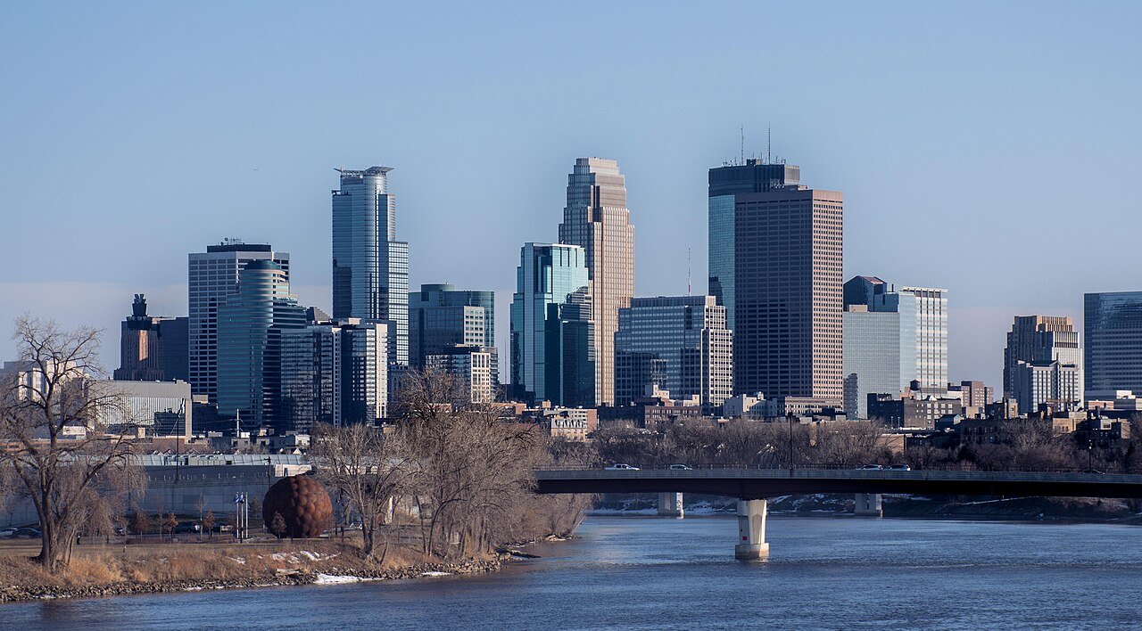 Skyscrapers in a city skyline viewed from across a river, with a bridge and leafless trees in the foreground.