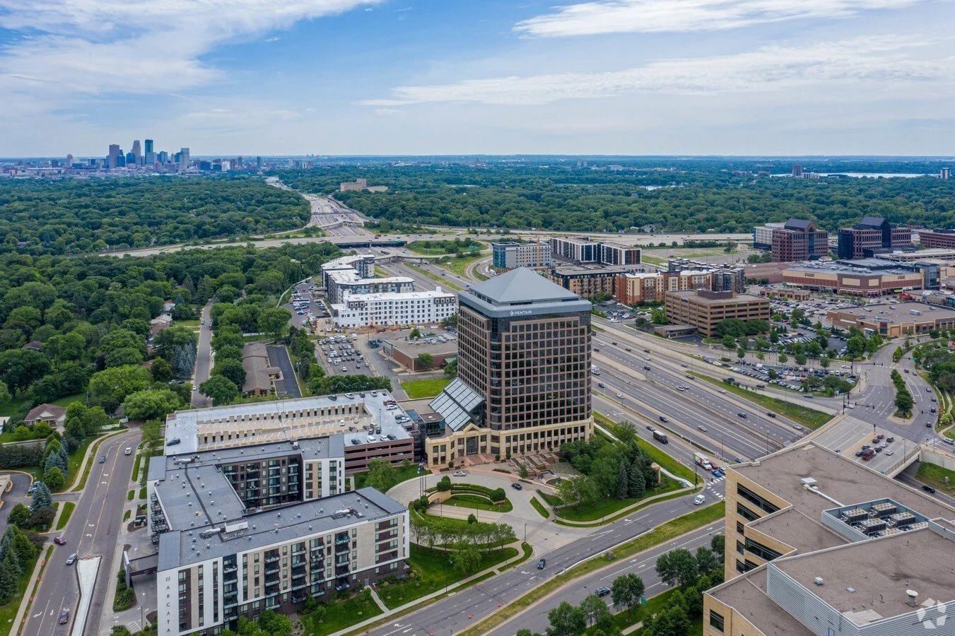 Aerial view of a cityscape with high-rise buildings, roads, and green parks, with a skyline in the distance under a partly cloudy sky.
