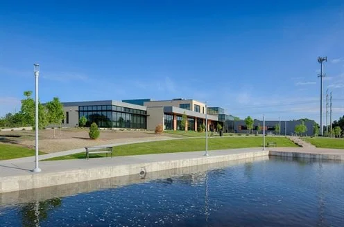 Modern building along a waterfront with a concrete walkway, lampposts, and a bench under a clear blue sky.