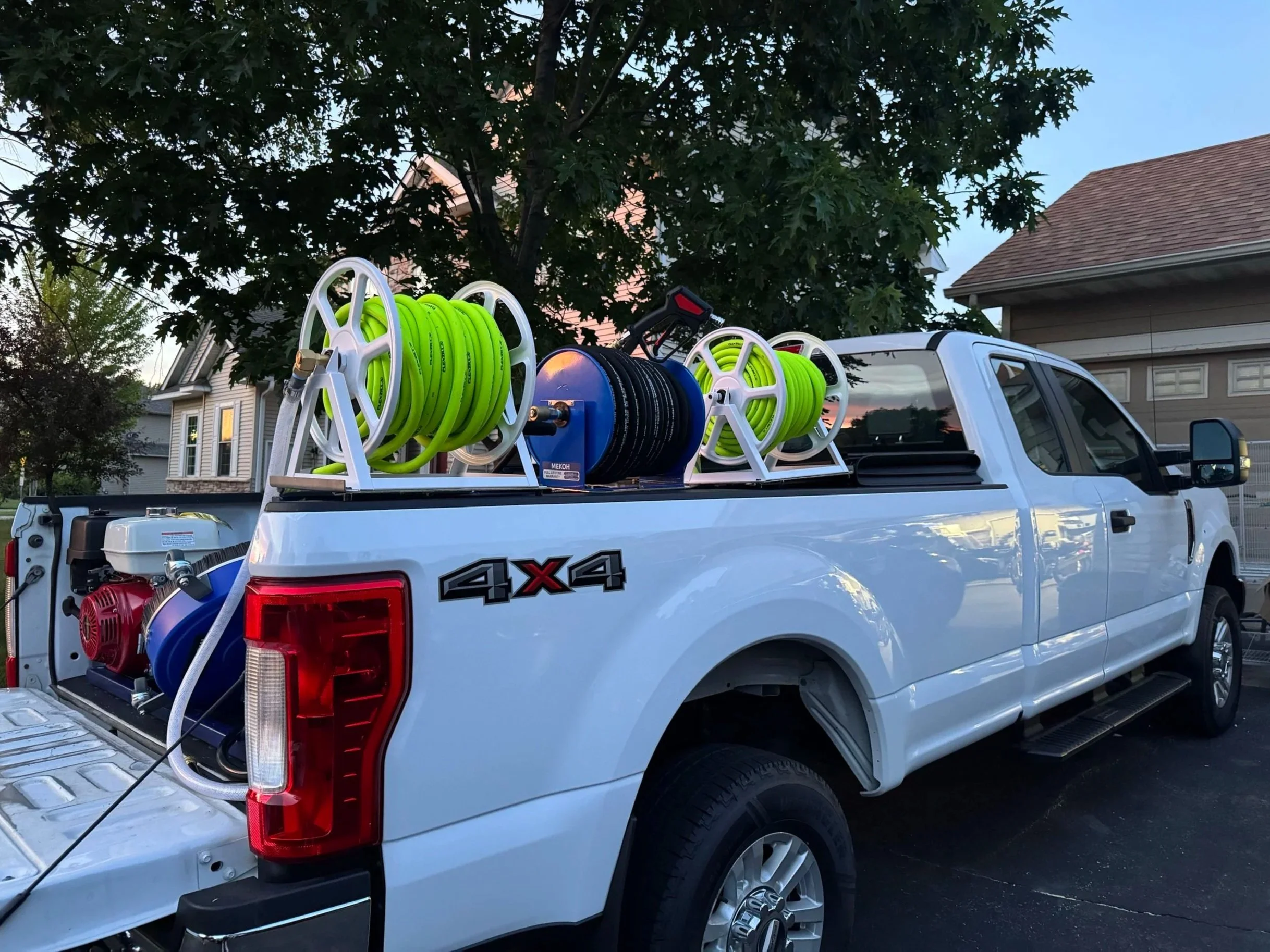 A white pickup truck with a ladder rack carrying bright green and black hoses, parked in a residential neighborhood with houses, trees, and a blue sky in the background.
