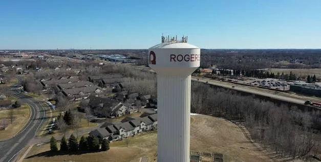 A tall white water tower with the Rogers logo and name on it, situated near a residential neighborhood and a highway.
