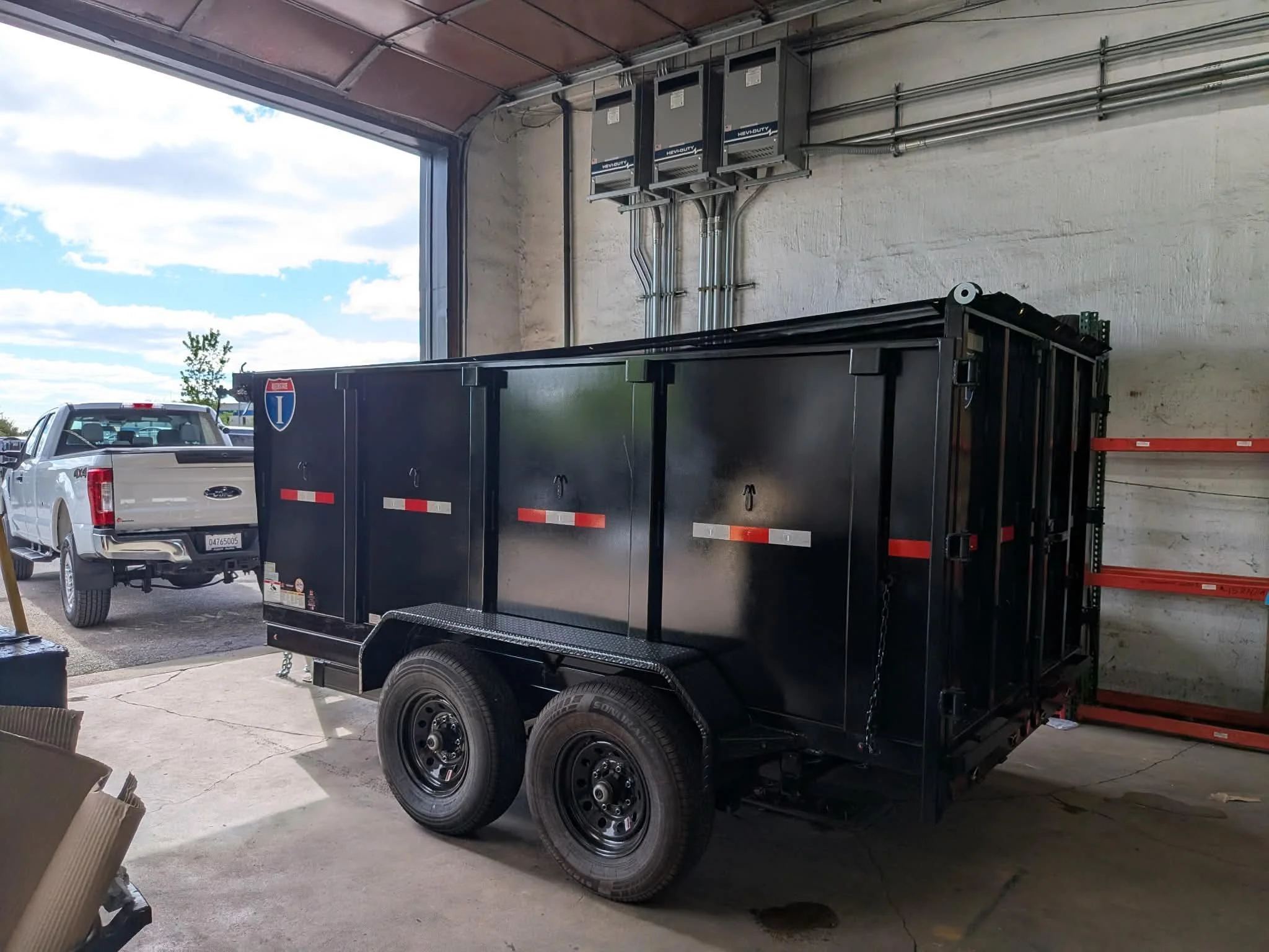 Black utility trailer parked inside a garage, with a silver pickup truck outside and cloudy sky visible through the open garage door.