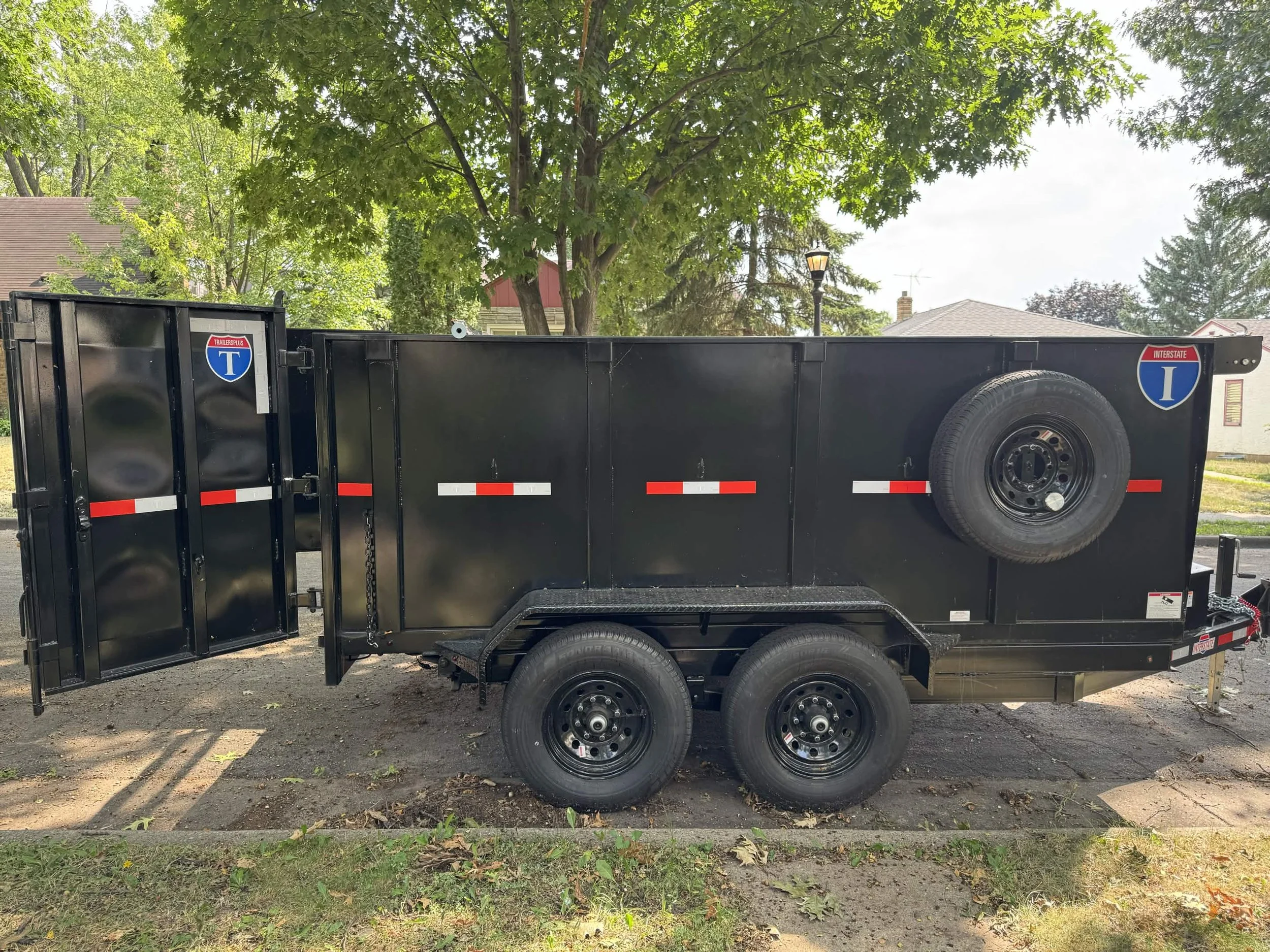 A black utility trailer with a large wheel in the middle and open fold-down sides, parked on a residential street with green trees, a house with siding, and a lamp post visible in the background.