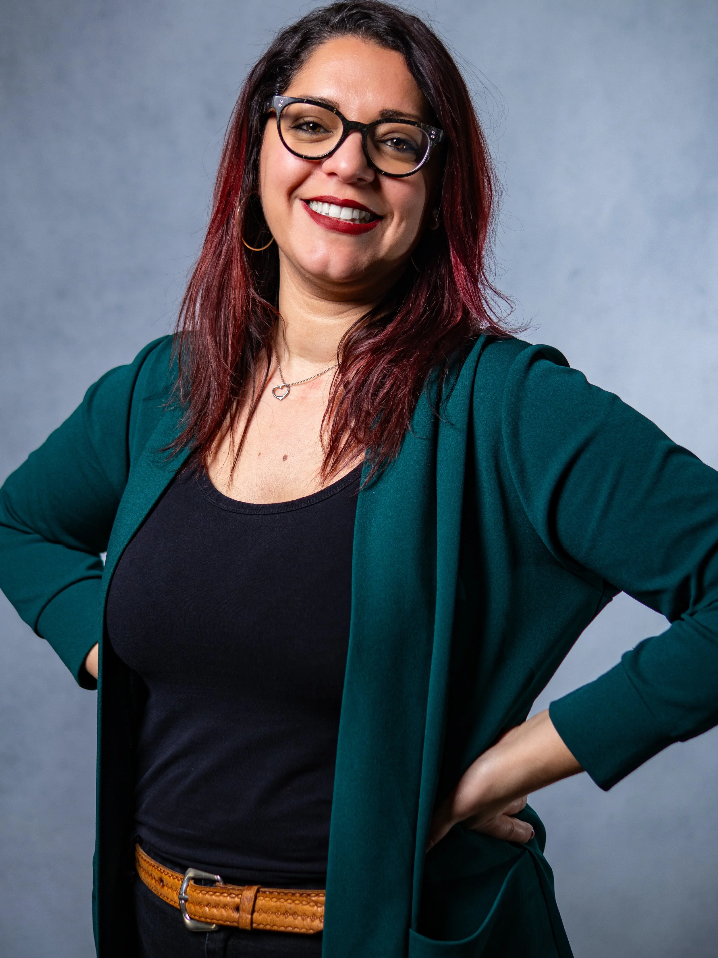 Une femme souriante avec des lunettes, portant un blazer vert foncé et un t-shirt noir, avec un collier en forme de cœur, posant avec la main sur la hanche contre un fond gris.
