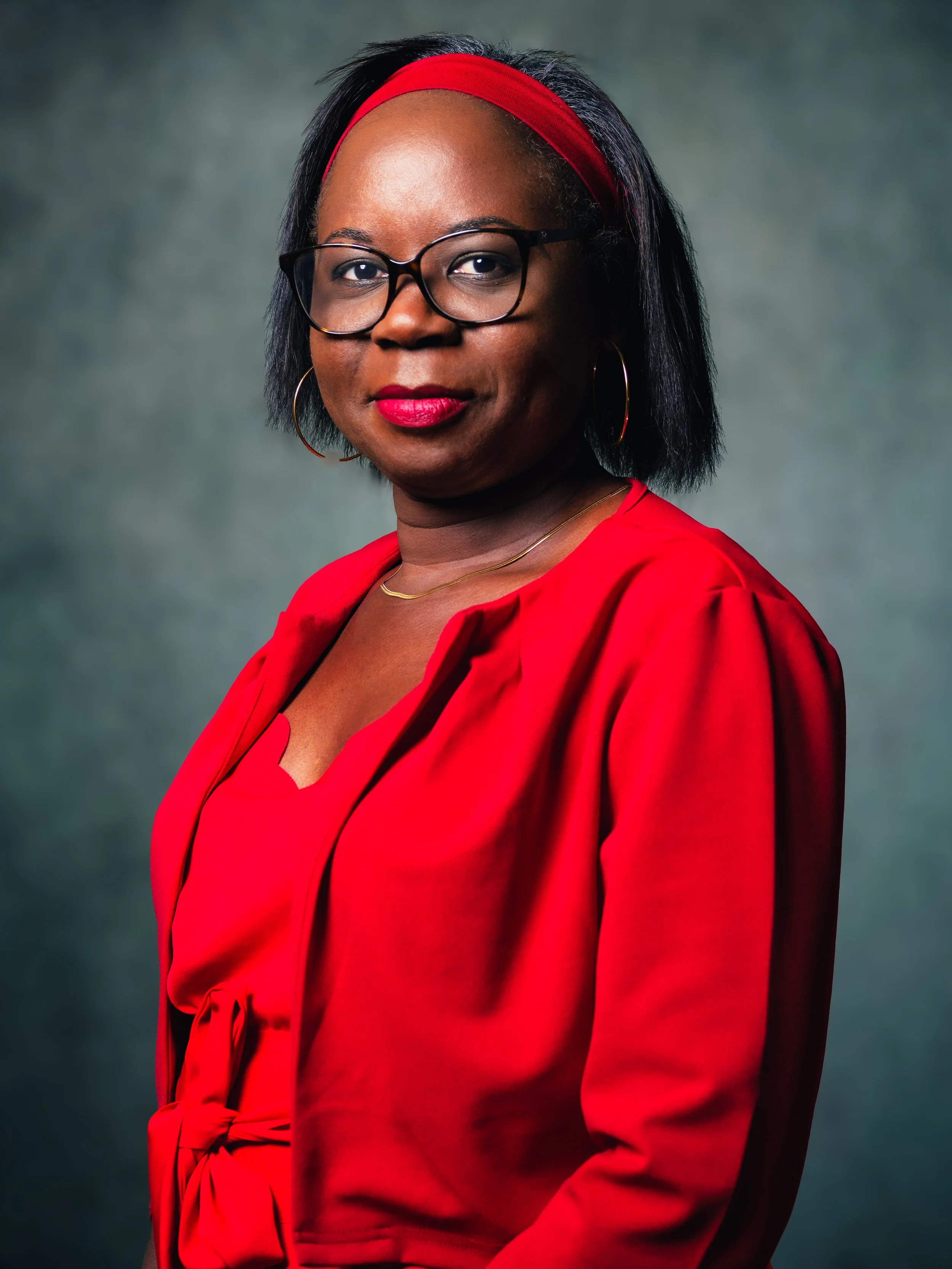 Portrait d'une femme portant des lunettes, un chemisier rouge et un bandeau rouge, avec un fond flou de couleur gris-vert.