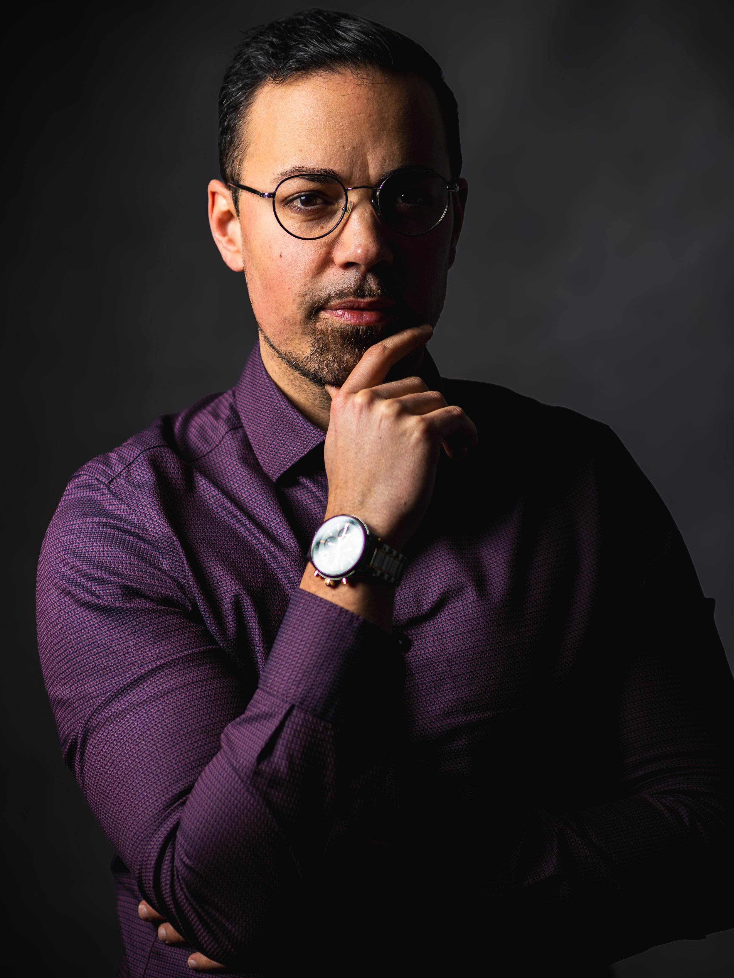 Portrait d'un homme avec lunettes, en profonde réflexion, portant une chemise violette à petits motifs et une montre élégante, devant un fond sombre.