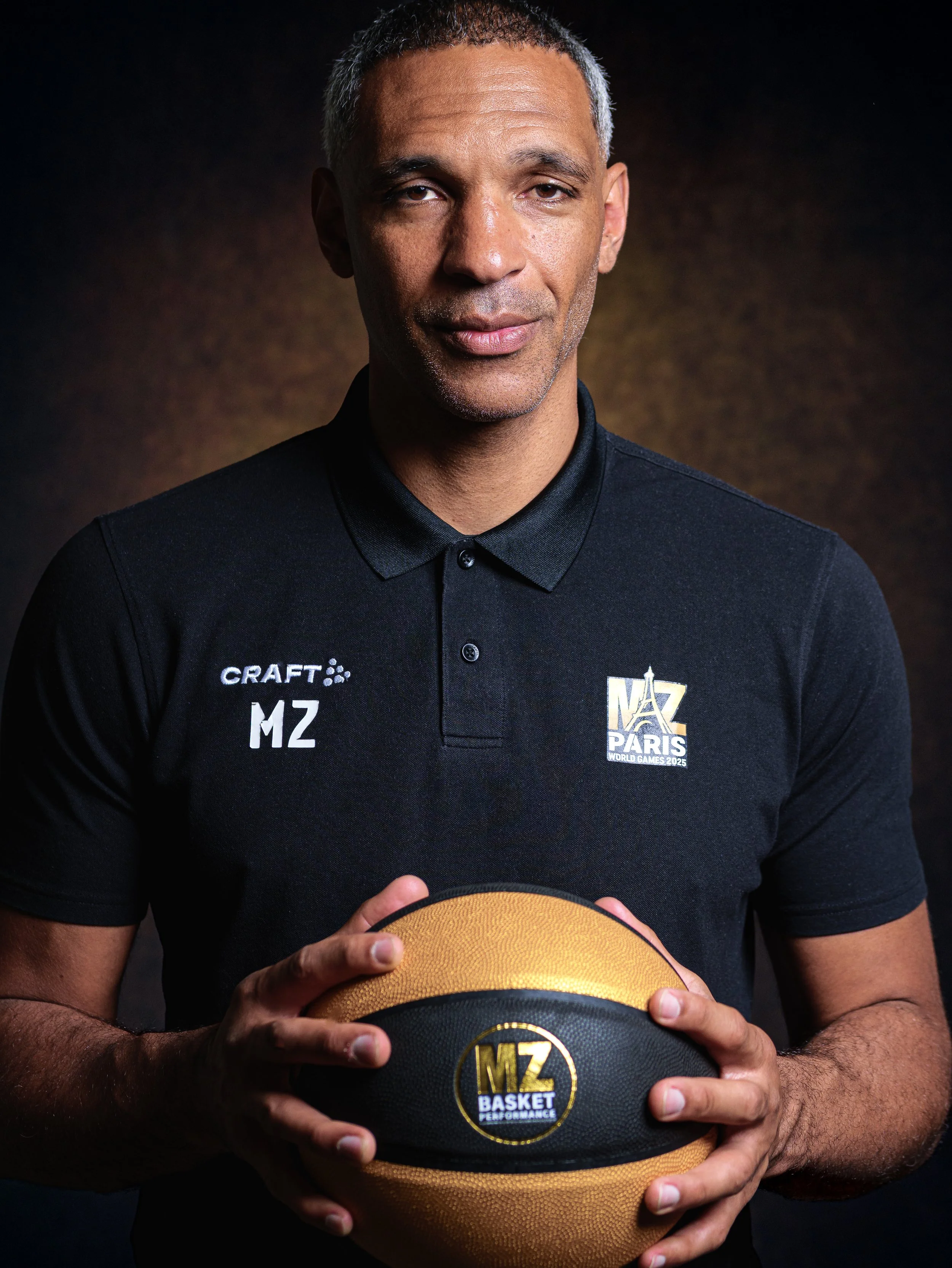 Un homme tenant un ballon de basket-ball, portant un polo noir avec le logo 'MZ PARIS' et 'WORLD GAMES 2025'.