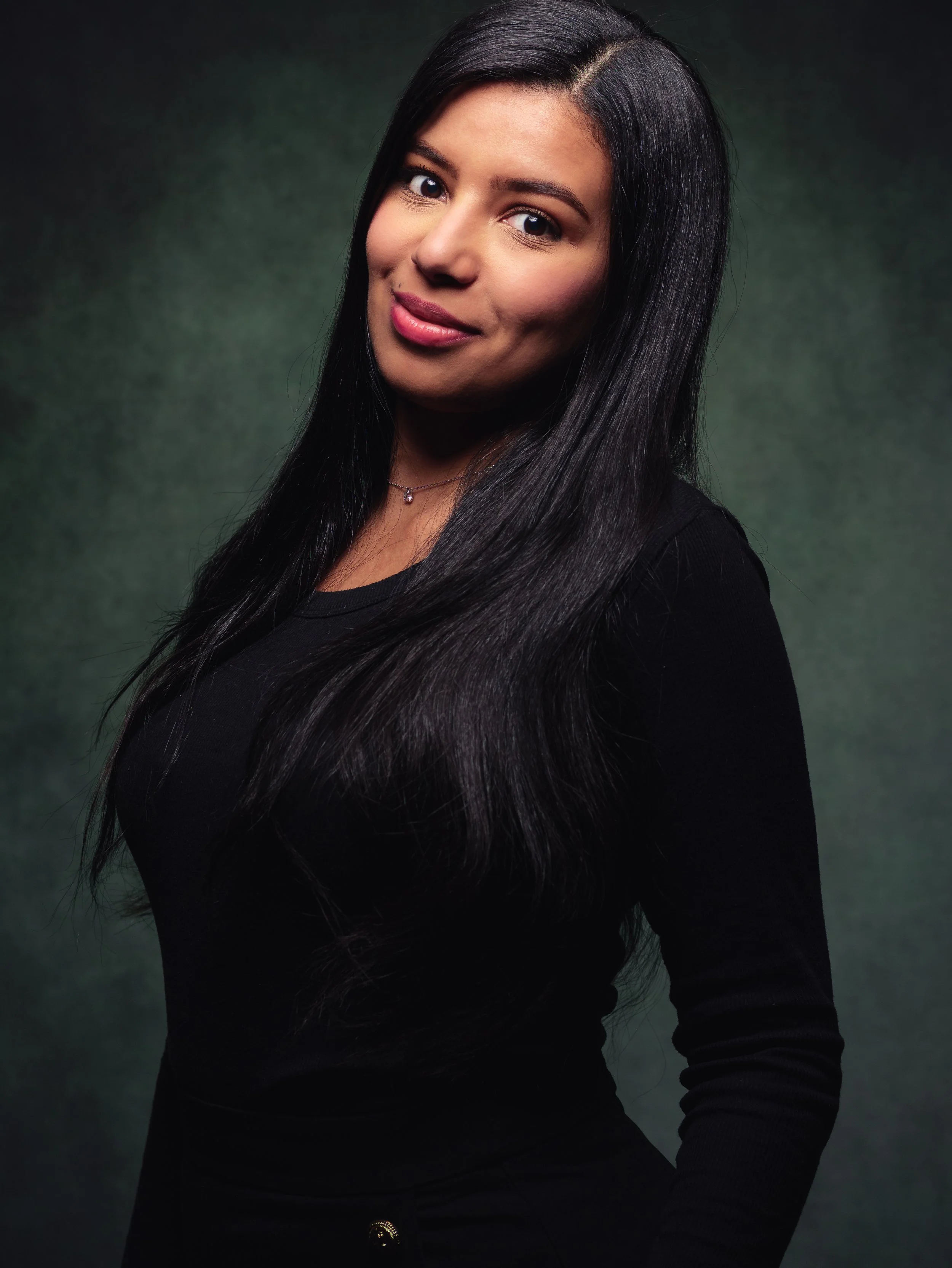 Portrait de femme avec long cheveux noirs, portant un haut noir et un collier, contre un fond sombre et vert.