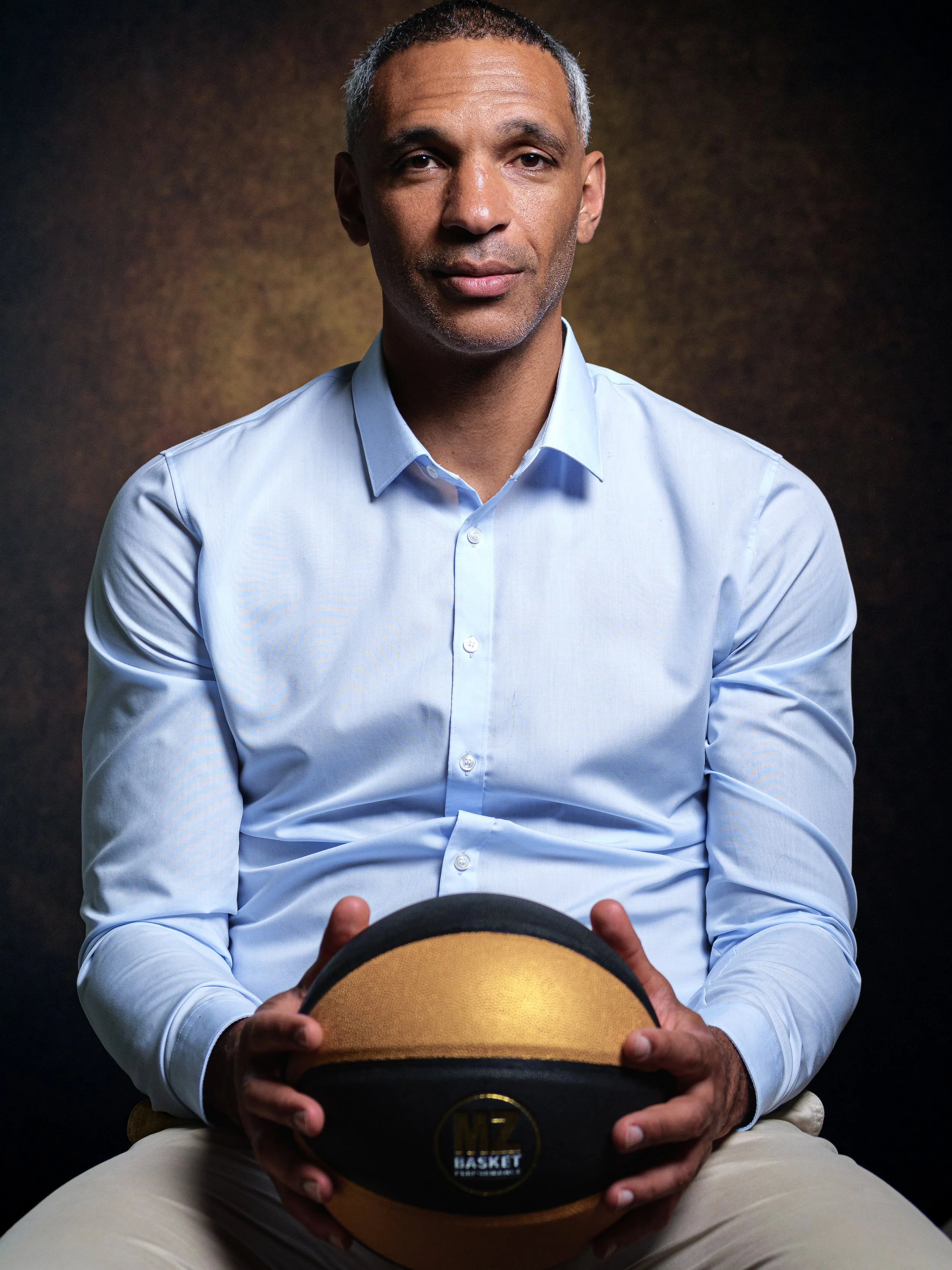 Un homme assis, portant une chemise bleue et tenant un chapeau de basketball.