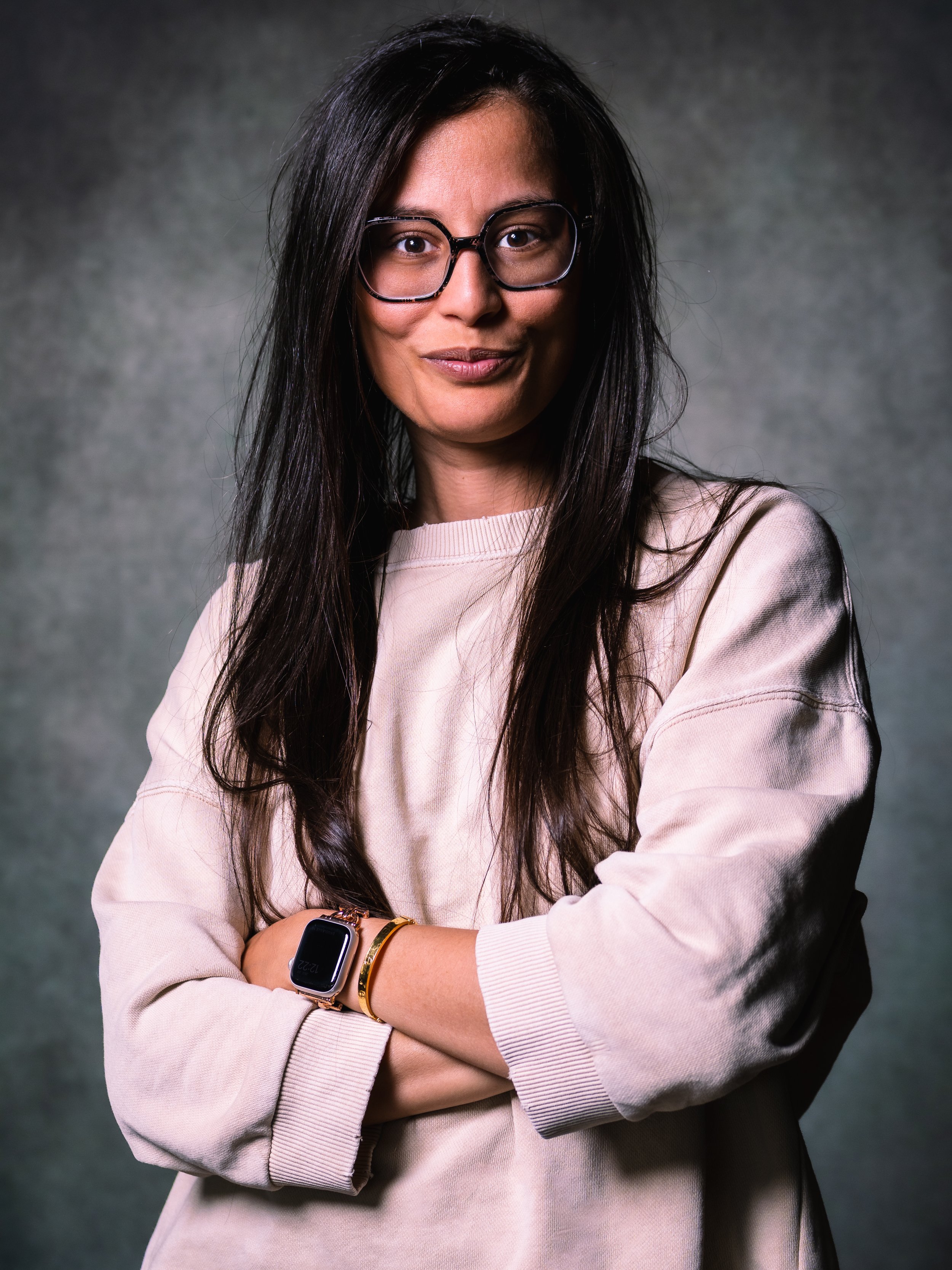 Jeune femme avec des lunettes noires, cheveux longs et noirs, portant un sweat-shirt beige avec des bracelets, un smartwatch, et une expression confiante, croisée bras, contre un fond gris.