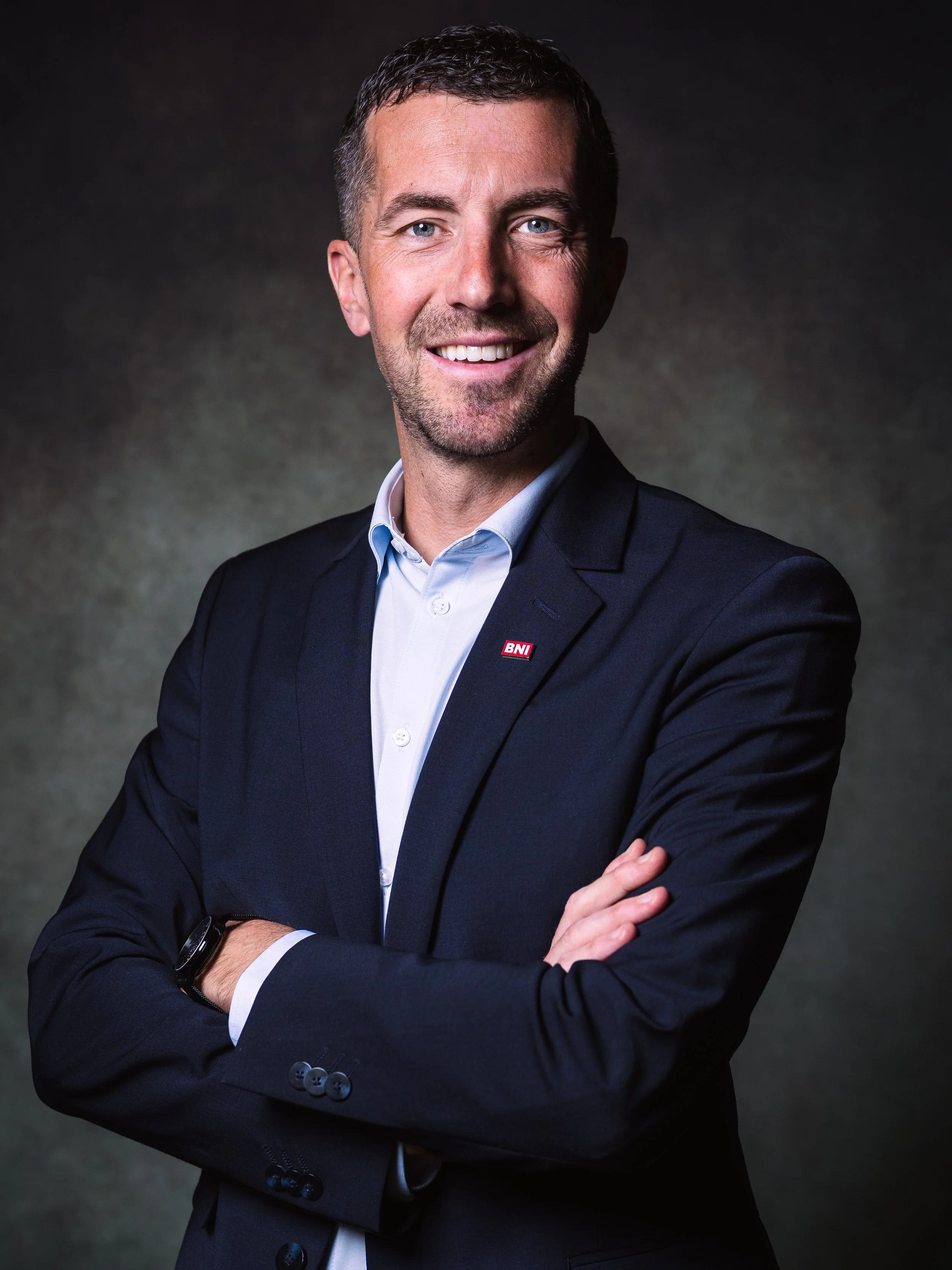 Homme d'affaires souriant portant un costume noir, une chemise blanche et une montre noire, posé contre un fond sombre.