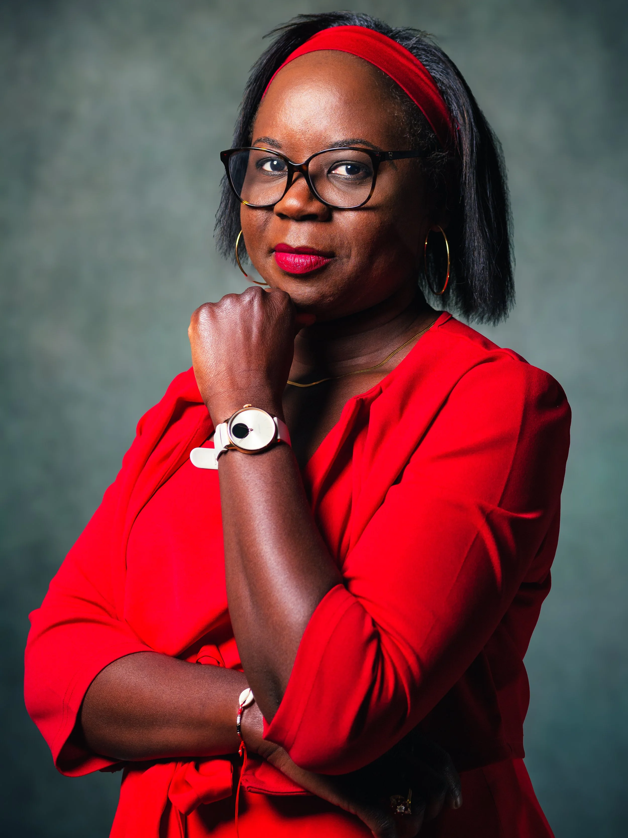Portrait d'une femme noire portant des lunettes, un bandeau rouge et une robe rouge, posant avec le bras croisé et la main sous le menton contre un fond flou.
