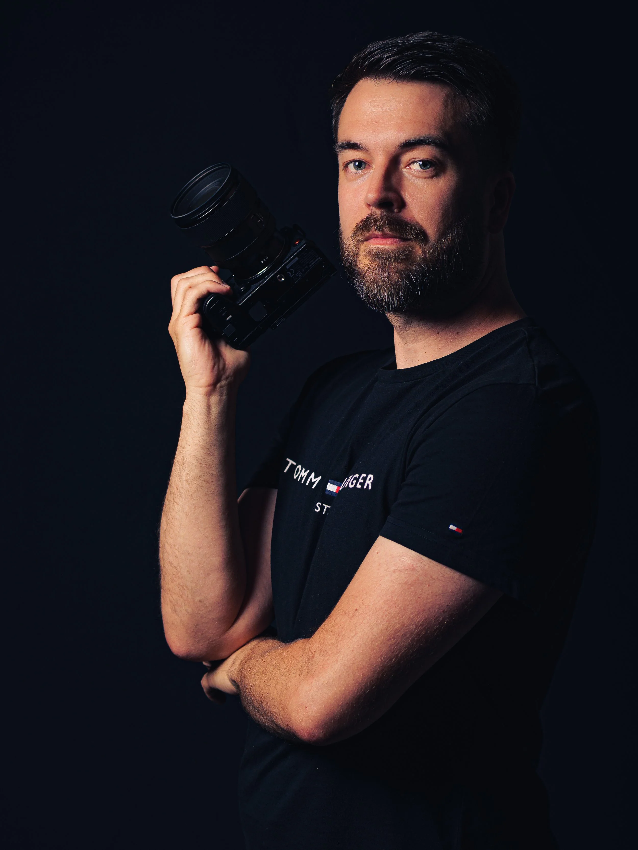 Homme avec une barbe, portant un t-shirt noir, tenant un appareil photo avec objectif.