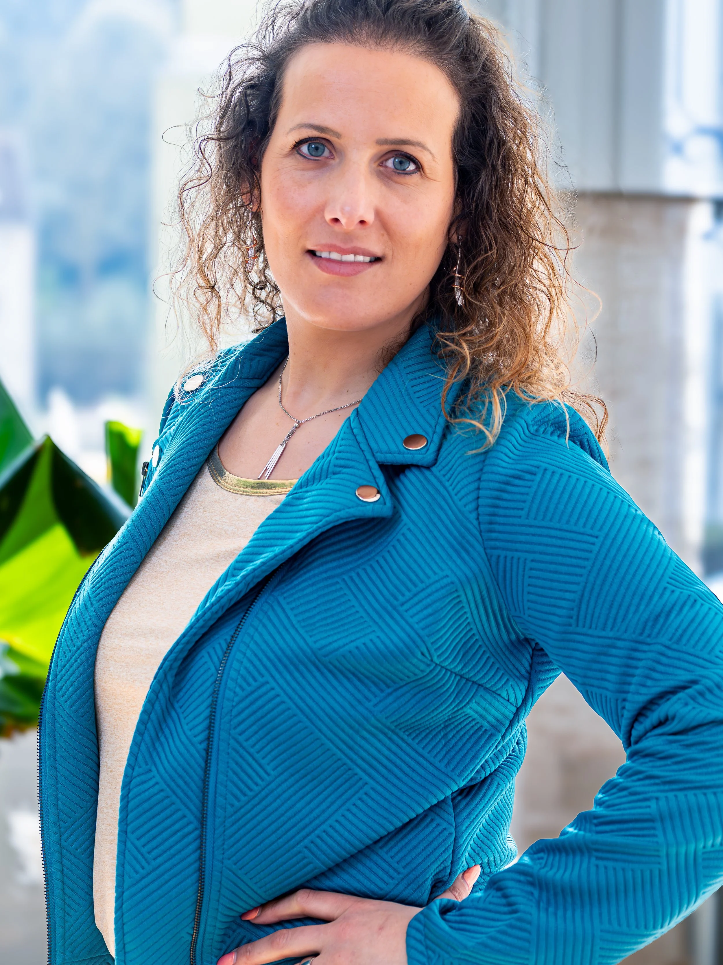Une femme aux cheveux bouclés, portant une veste bleue et un haut beige, pose dans un environnement intérieur lumineux avec des plantes en arrière-plan.
