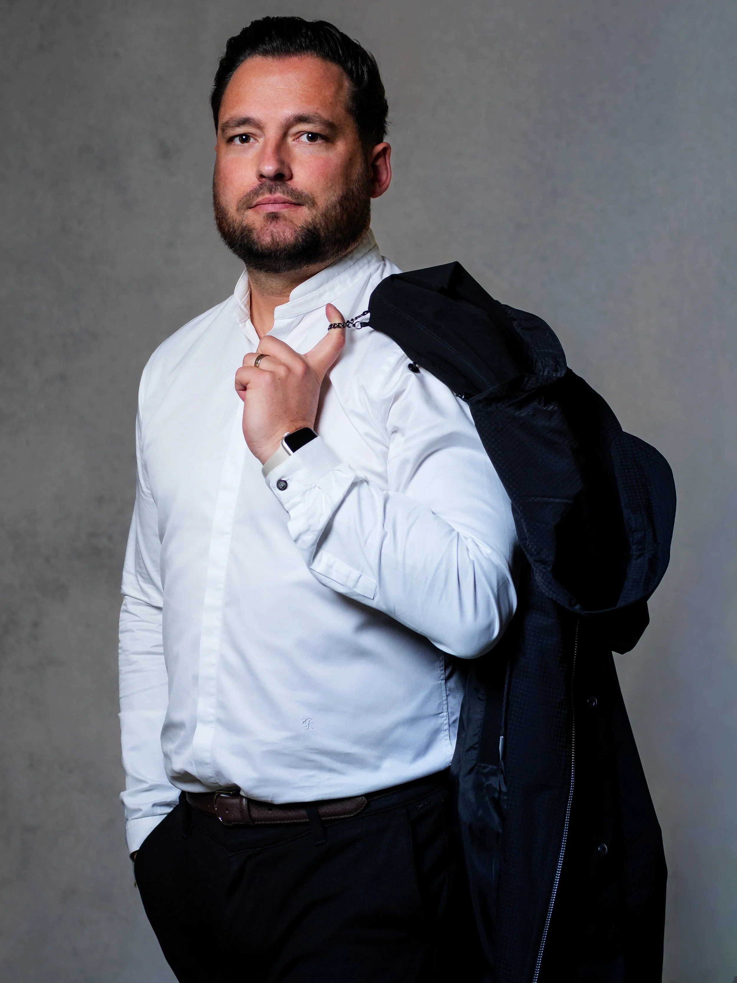 Un homme portant une chemise blanche, tenant une sacoche sur son épaule gauche, porte une montre intelligente au poignet gauche, pose contre un fond gris, regarde droit devant.