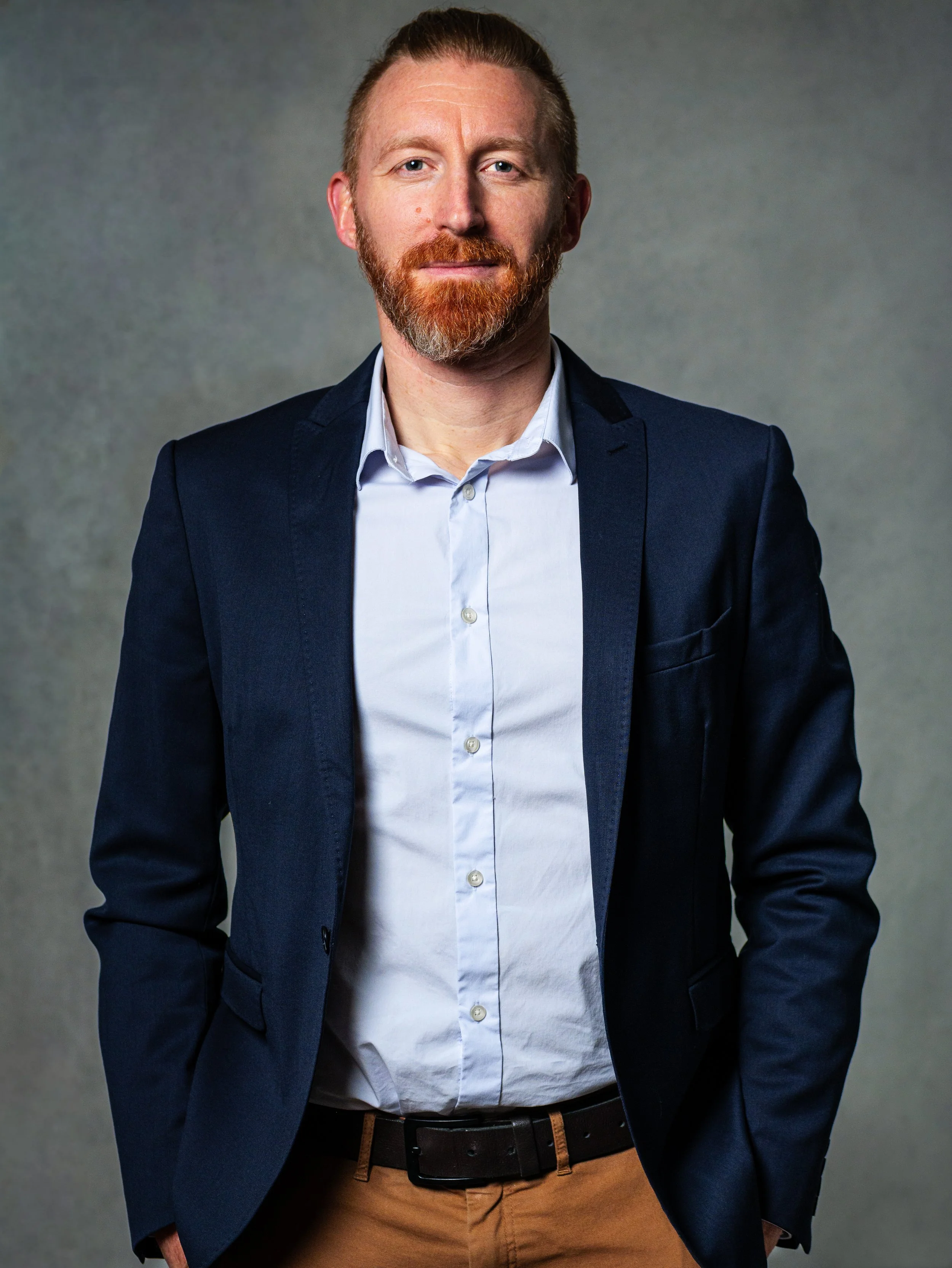 Homme d'affaires avec barbe rousse, porte une veste bleu marine et une chemise blanche, pose avec les mains dans les poches, fond gris