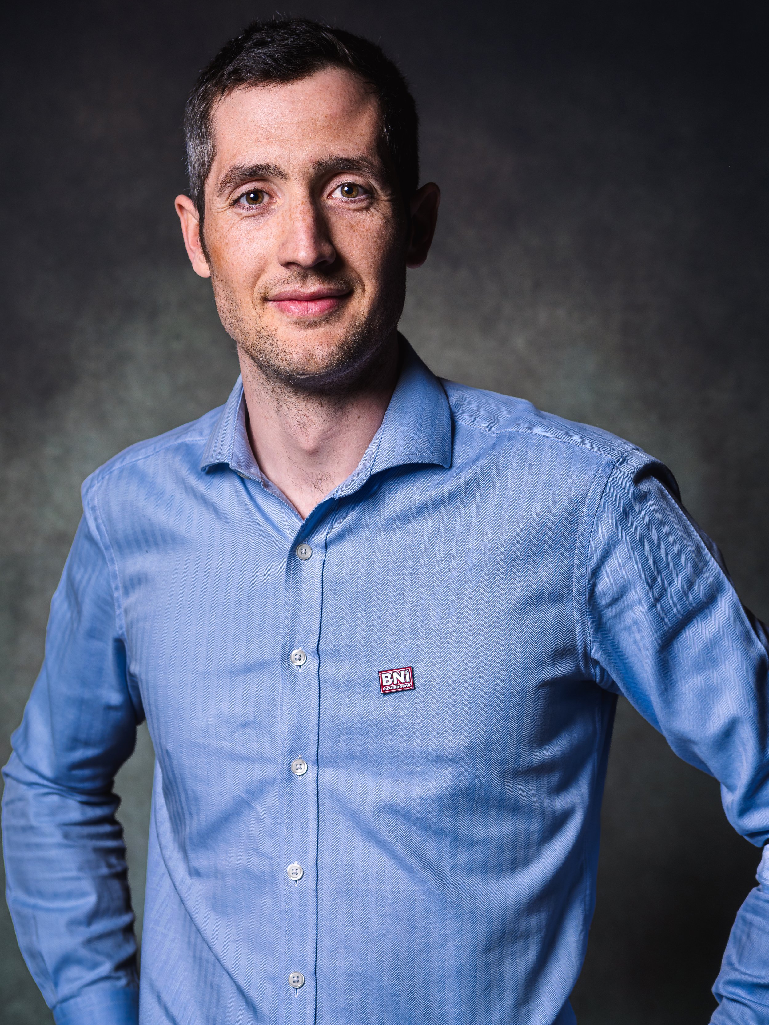 Homme en chemise bleue avec un badge 'BNI' sur la poitrine, pose sur fond sombre, regardant l'objectif, sourire subtil.