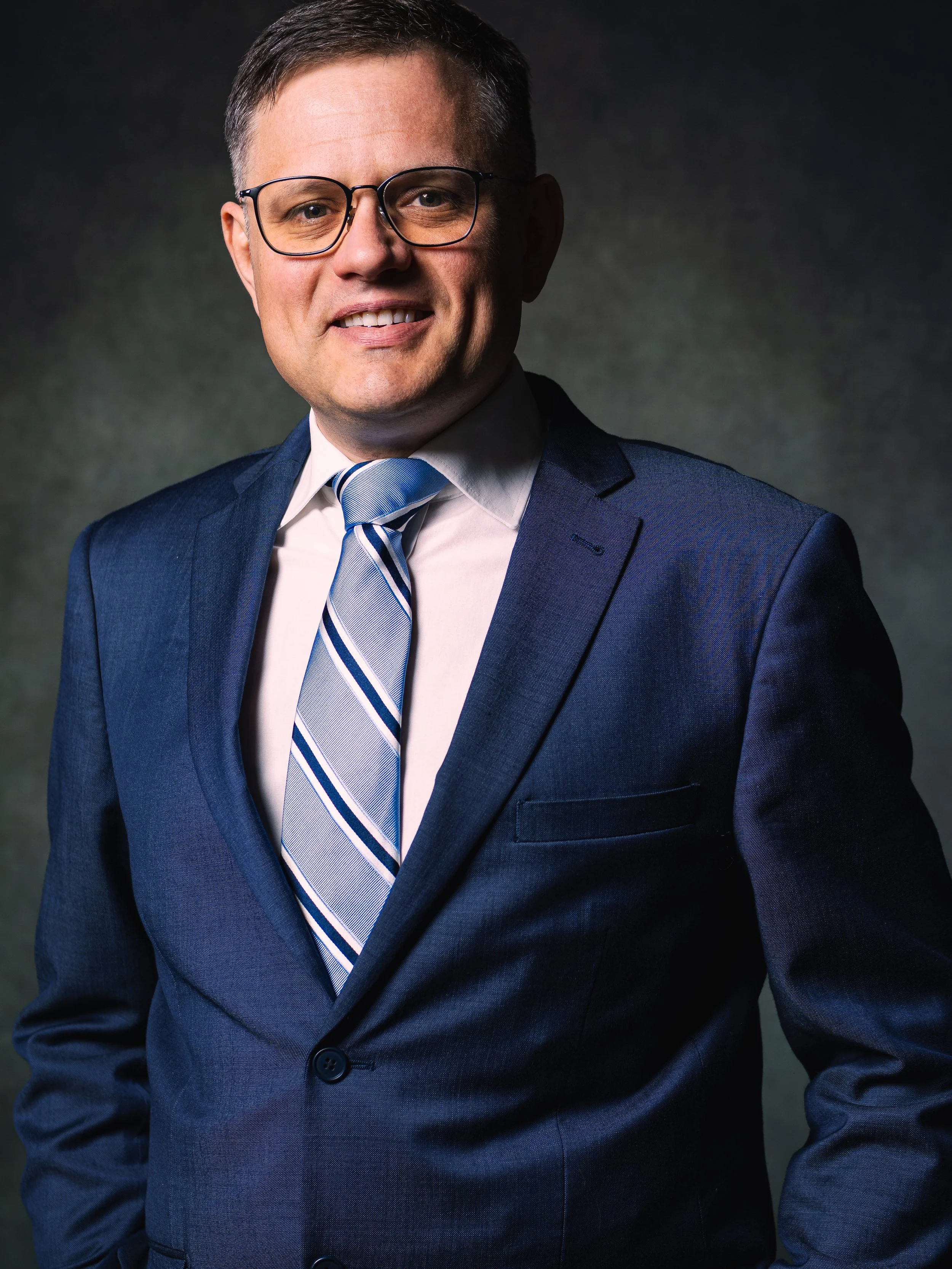 Portrait d'un homme d'affaires portant un costume bleu, une chemise blanche et une cravate rayée, souriant légèrement, avec des lunettes, sur un fond sombre.