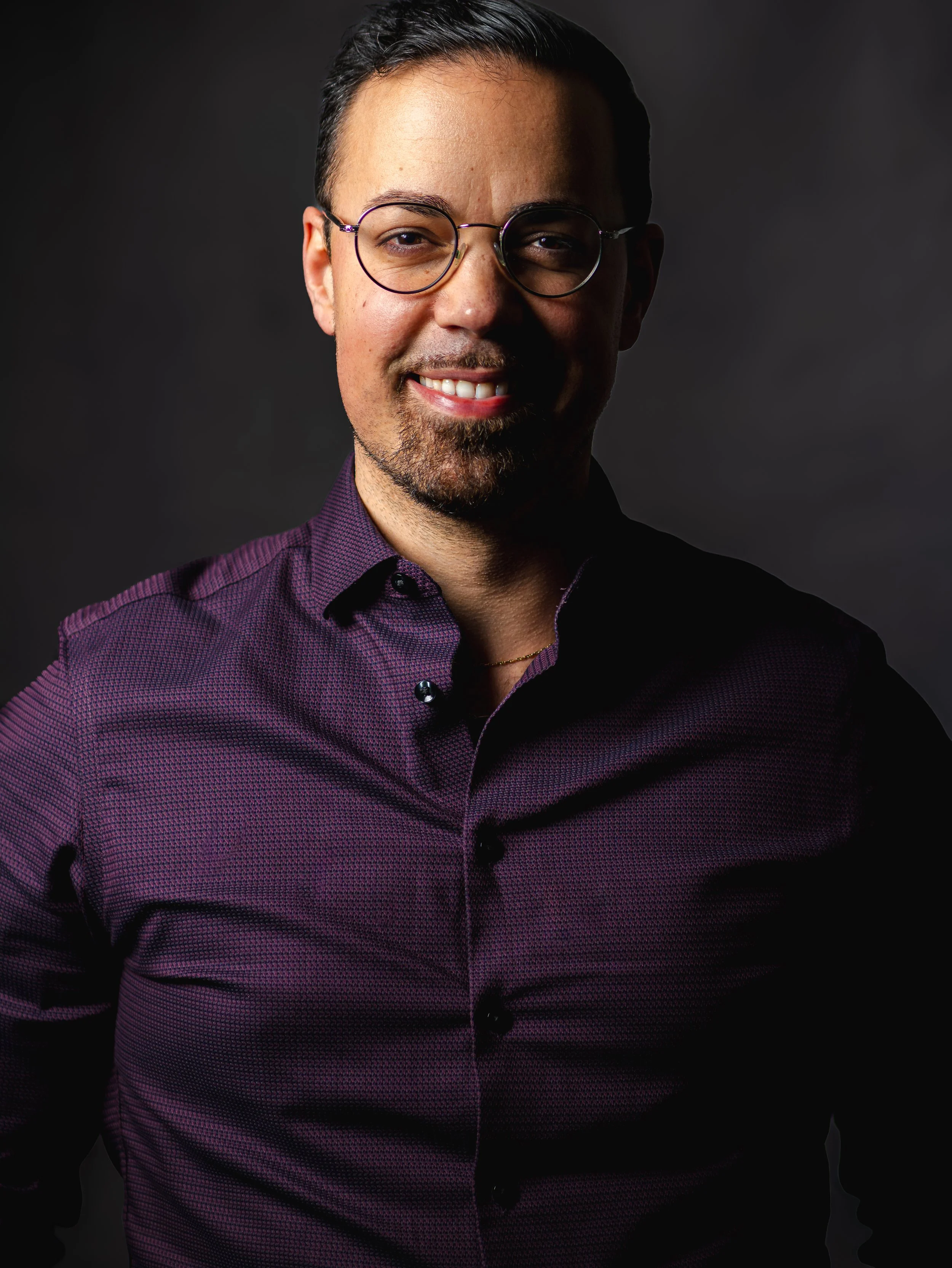 Portrait d'un homme souriant portant une chemise à manches longues violette et des lunettes rondes, avec un fond sombre.