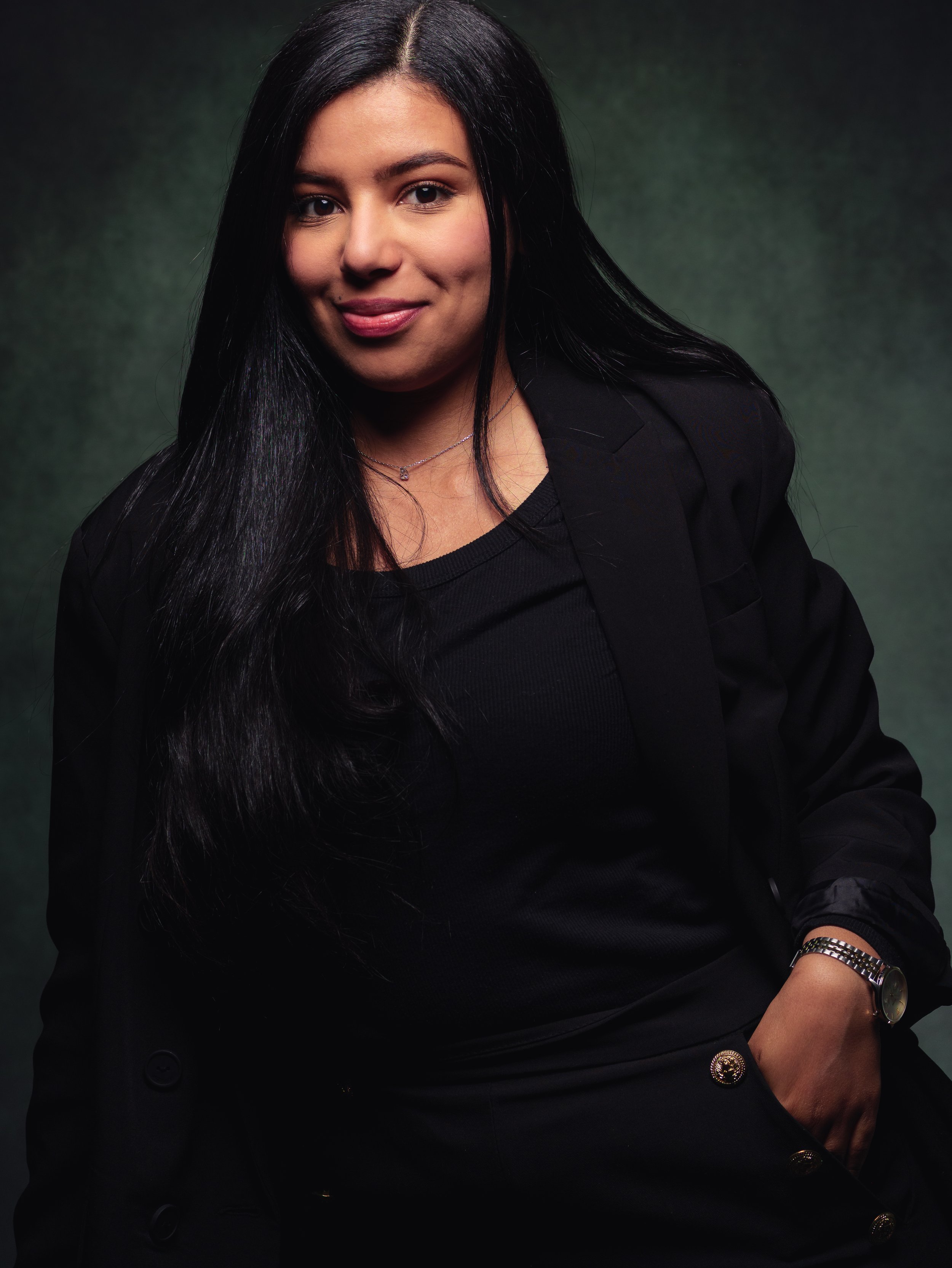 Une femme aux cheveux longs et noirs, portant un blazer noir et un collier, pose contre un fond sombre.