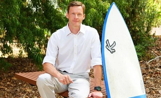 Man sitting on a park bench next to a white surfboard with blue trim, outdoors with green trees in the background.