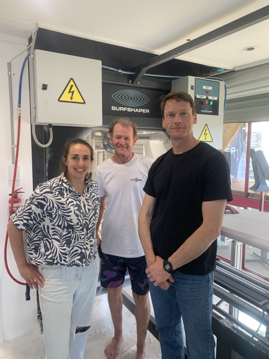 Three people standing inside a facility with machinery, including a woman wearing a patterned blouse and two men, one of whom is barefoot, and the machine labeled 'Surfshaper' in the background.