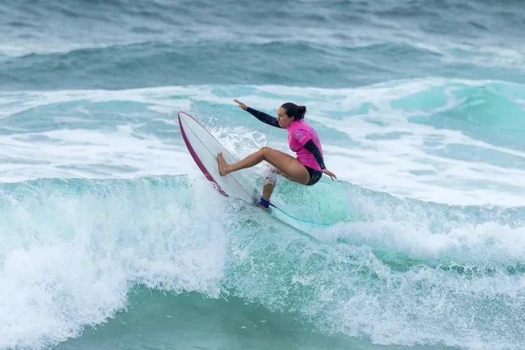 A woman surfing on a wave, balancing on her surfboard, wearing a pink and black wetsuit with a pink vest.