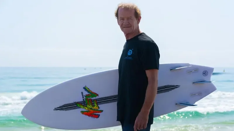 A man standing on the beach holding a surfboard with a graphic design while smiling at the camera, ocean waves in the background.