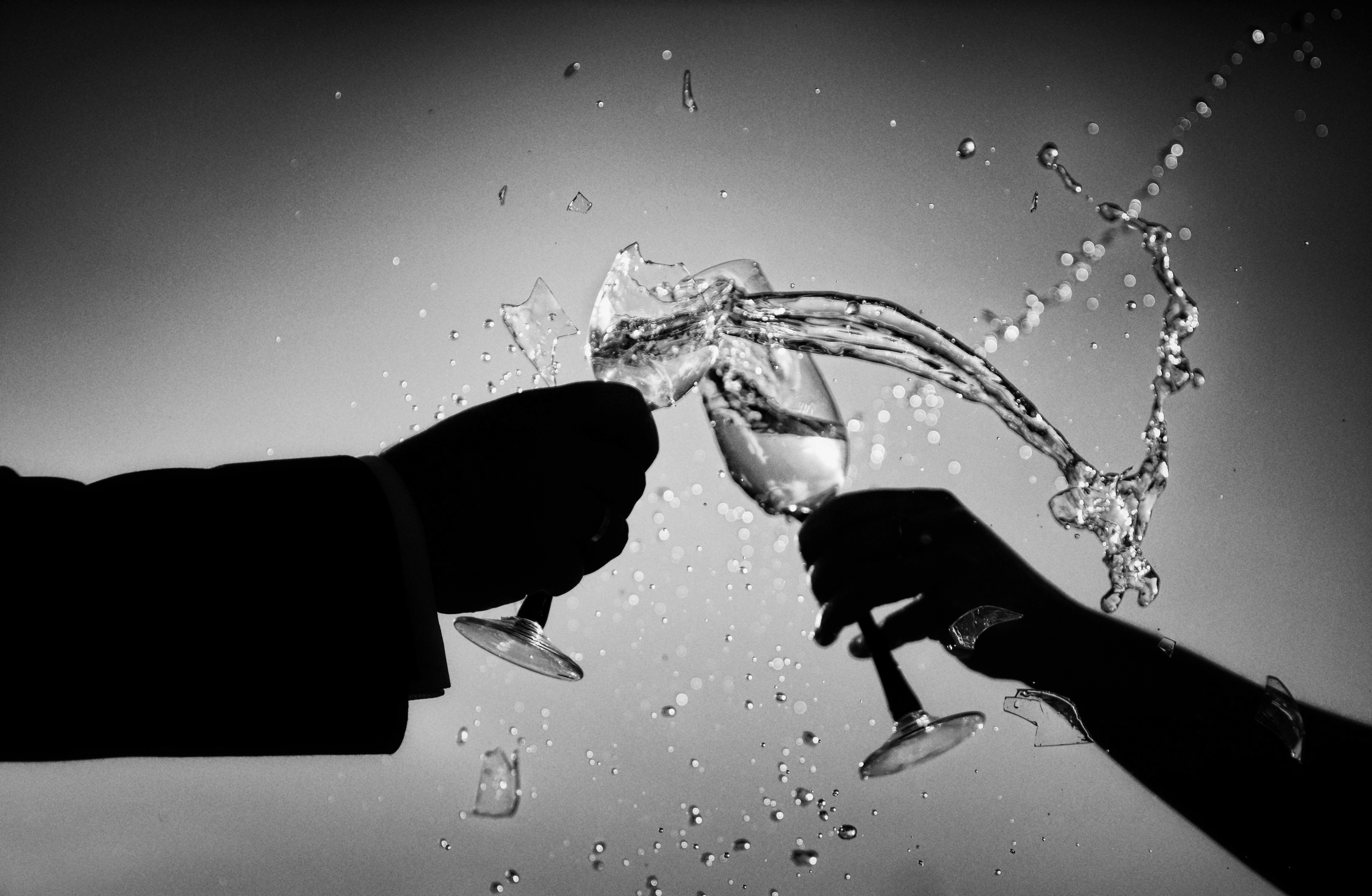 Silhouette of two people clinking glasses with shattered glass and water splashing against a gray background.