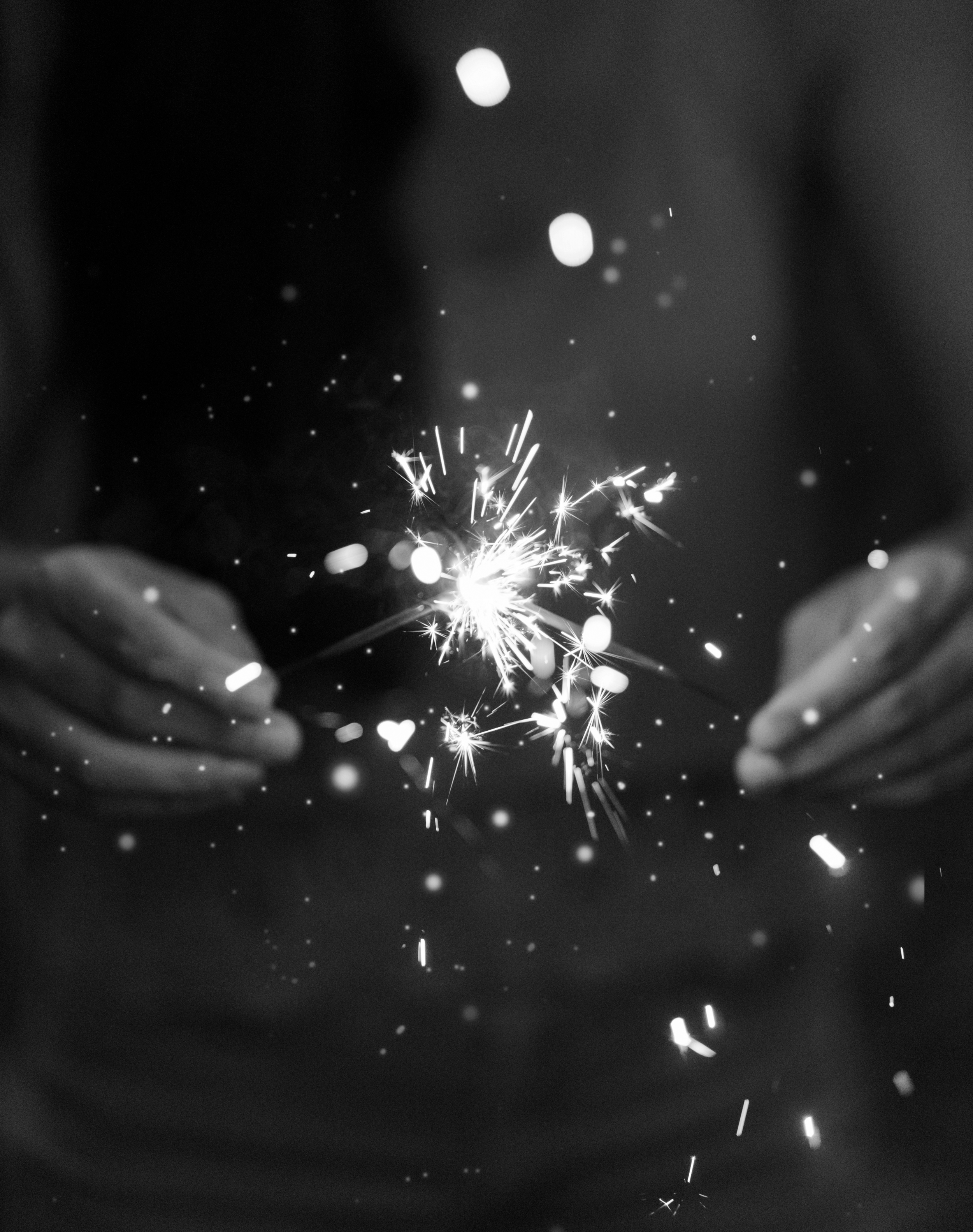 Black and white photo of a hand holding a sparkler, with sparks flying and a dark background.