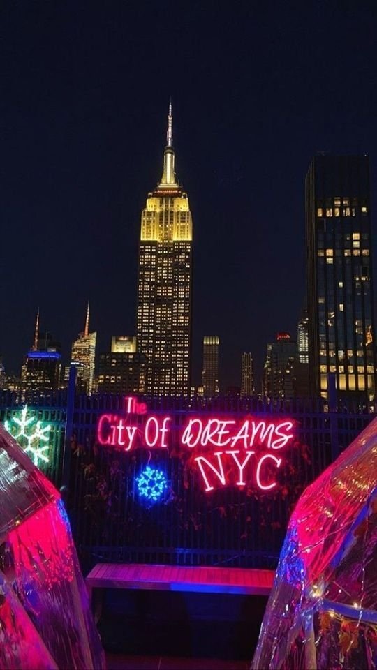 Nighttime view of New York City with illuminated Empire State Building and colorful neon sign that reads 'The City Of D R E A M S NYC' with decorative snowflake and lights.