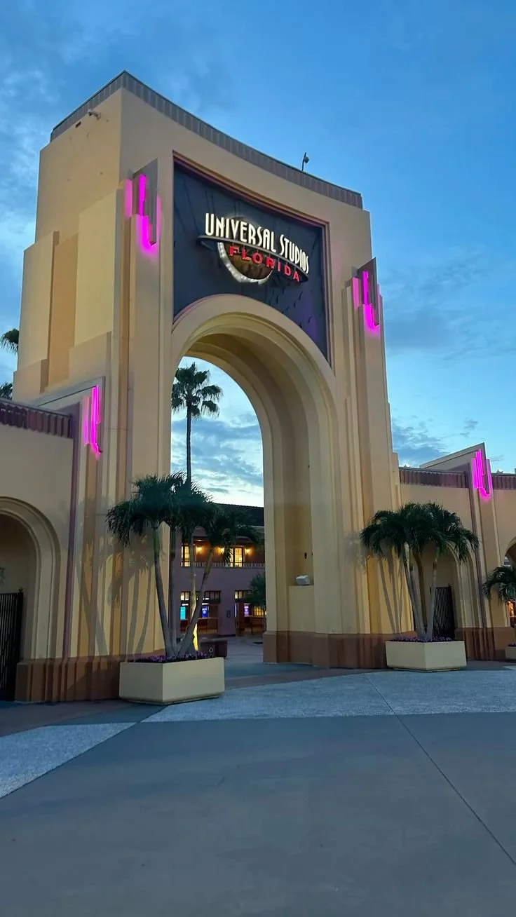 Entrance archway of Universal Studios Florida at dusk, featuring a sign with the studio's logo and pink neon lights, palm trees, and a partly cloudy sky.