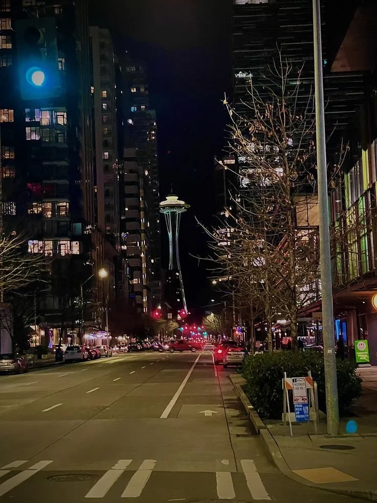 Nighttime city street scene with tall buildings, the Space Needle in Seattle in the background, traffic lights, parked cars, streetlights, and trees along the sidewalk.
