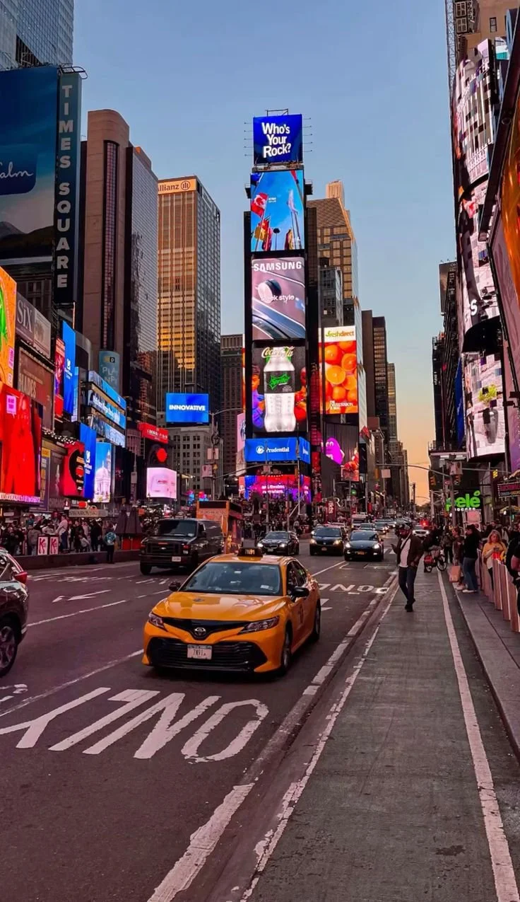 Time Square in New York City at dusk featuring a yellow taxi on the road, surrounded by bright billboards and skyscrapers with people walking on the sidewalk.