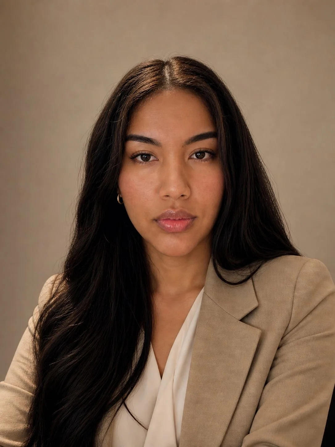 A young woman with long dark hair, wearing a beige blazer and tan top, looking into the camera with a neutral expression against a plain background.