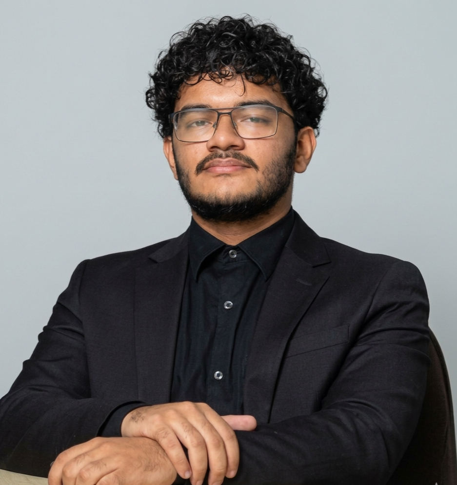Confident man with glasses, curly hair, and beard wearing a black suit and shirt, sitting with arms crossed against a plain gray background.
