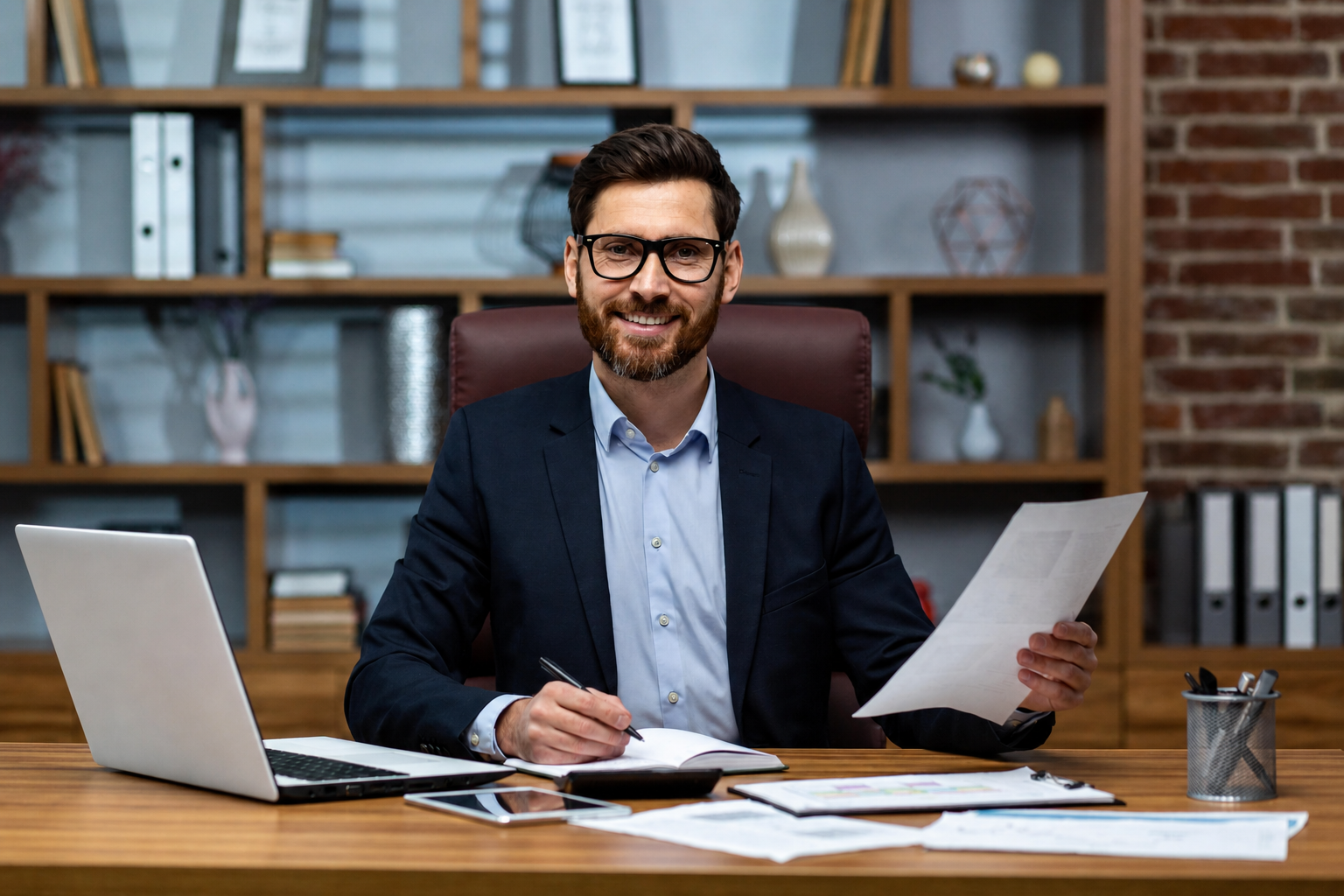 A smiling man with glasses and a beard sitting at a desk in an office, holding a sheet of paper, with a laptop and documents on the desk, in front of a bookshelf and brick wall.