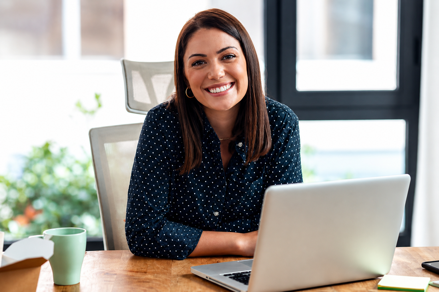 A woman with brown hair, wearing a navy blue polka dot shirt, smiling and sitting at a desk with a laptop in front of her, in a bright space with large windows.