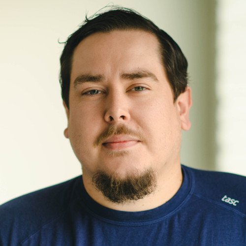 A young man with dark hair and a goatee, wearing a navy blue shirt, standing indoors near a window.