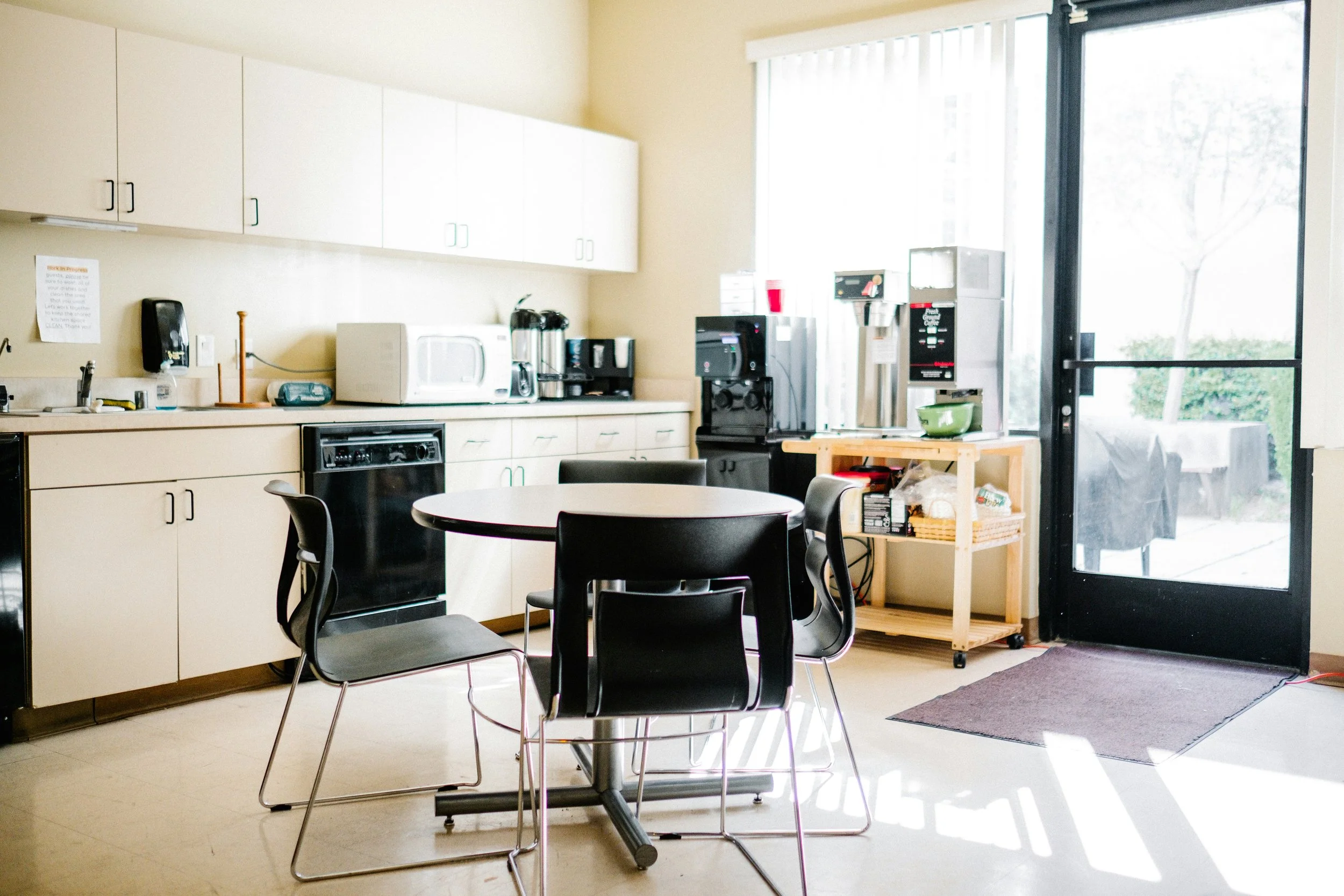 A kitchen with beige cabinets, a round black dining table with black chairs, and various appliances like a microwave, coffee maker, and a vending machine near a glass door. There is a small cart with supplies near the door and a sign posted on the wall.