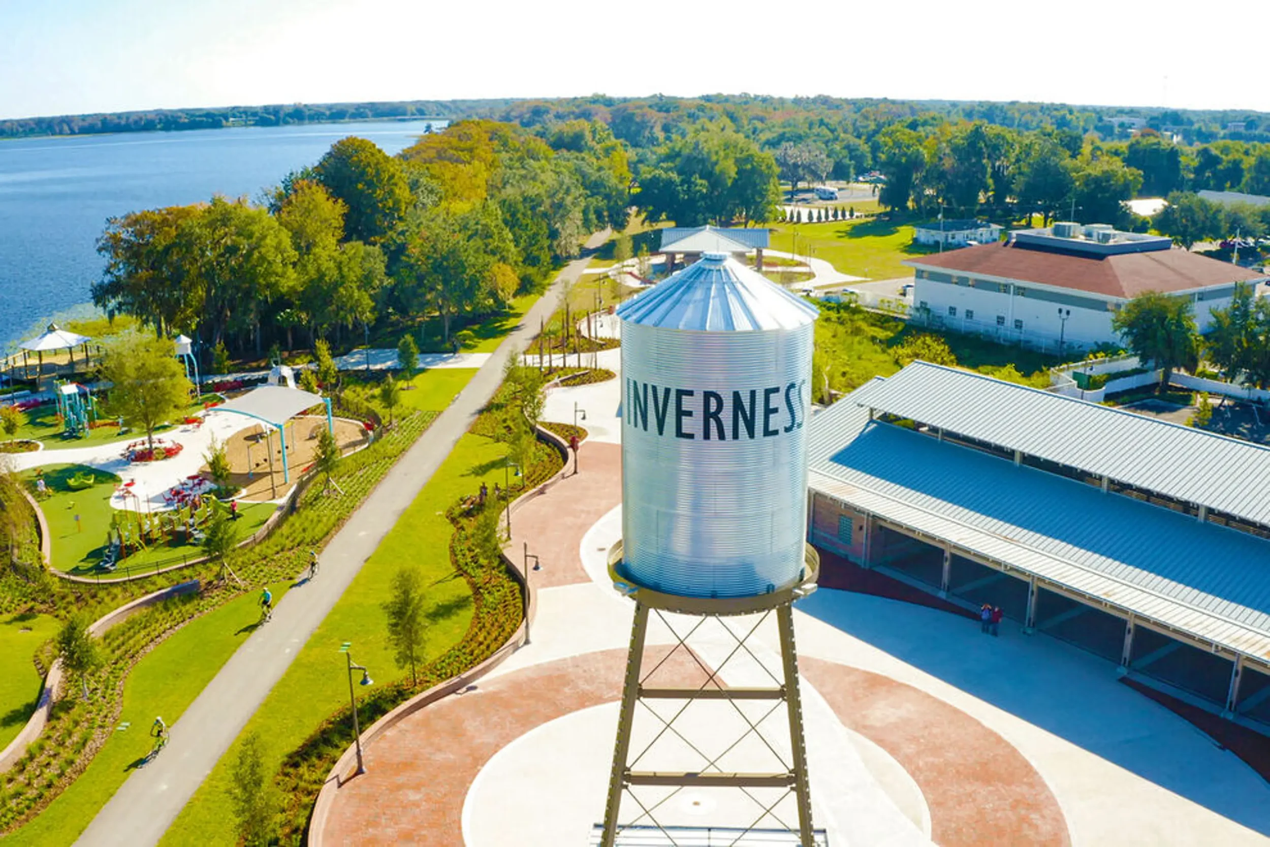 Aerial view of a park with trees, walking paths, playground equipment, and a large water tower with the word 'INVERNESS' on it, near a large body of water.