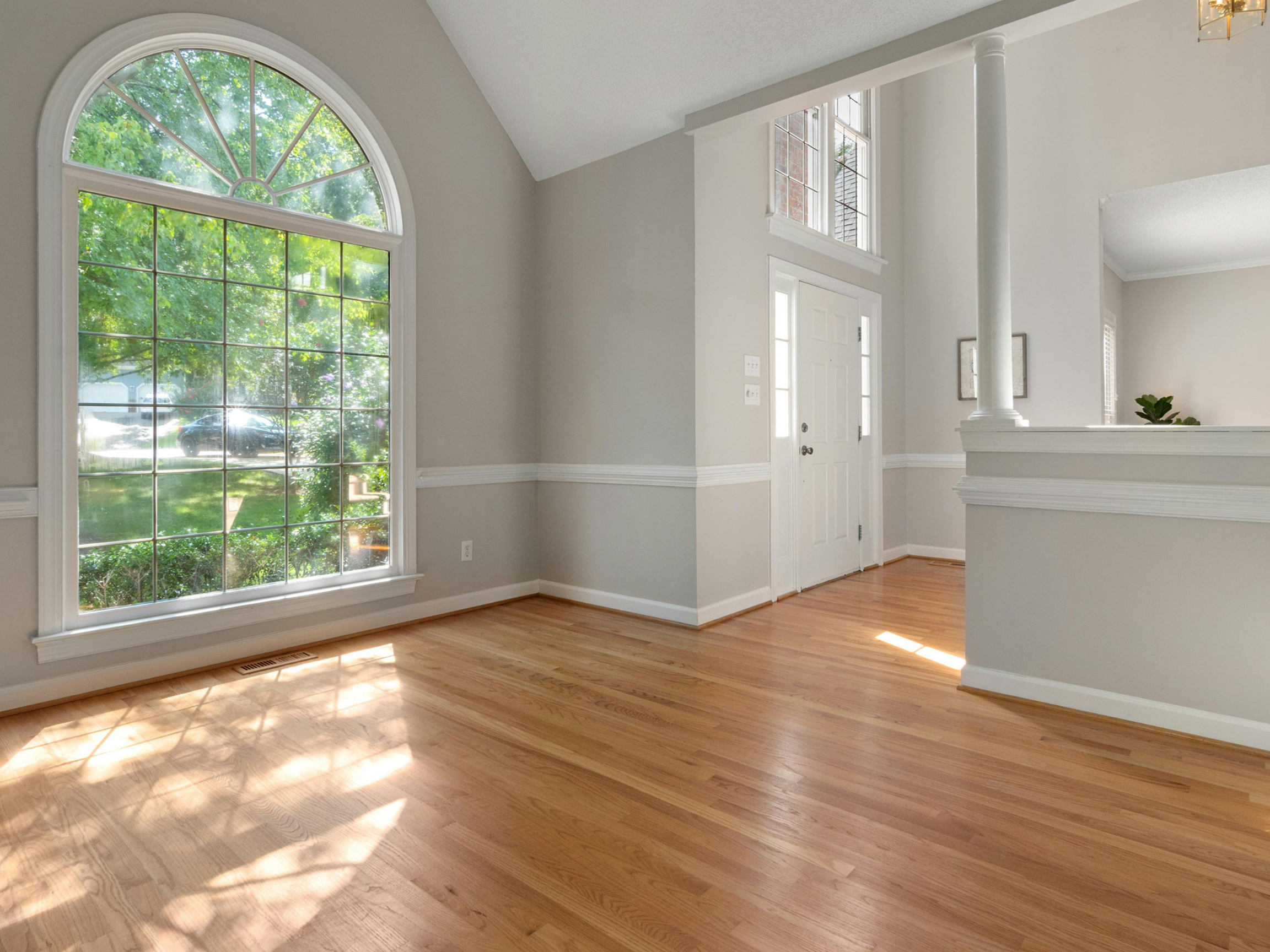 Interior of a bright living room featuring large arched and rectangular windows, light-colored walls, and hardwood floors.
