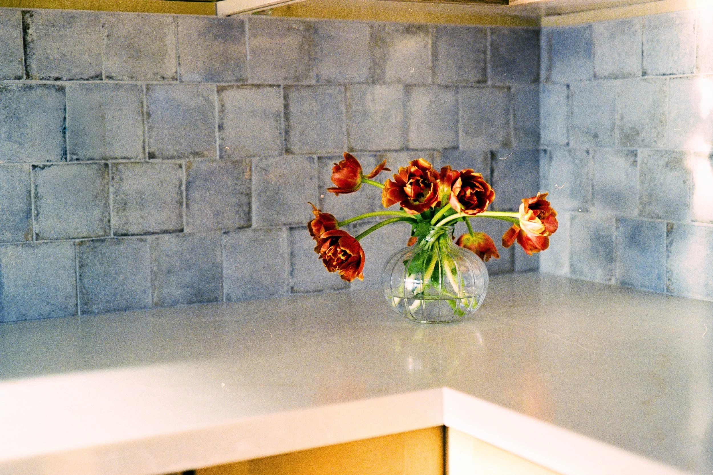 A clear glass vase with wilted red and orange tulips is placed on a white kitchen countertop against a gray brick wall background.