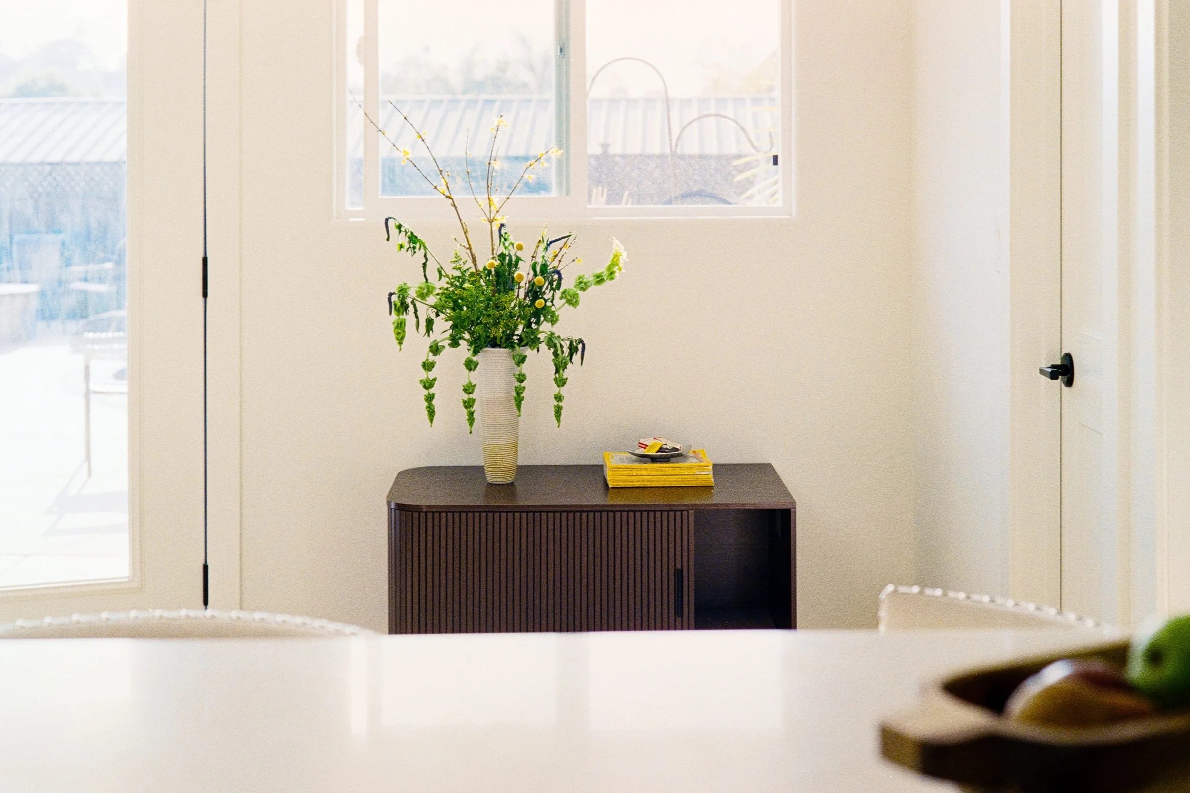 A vase with green and yellow flowers on a dark wooden table near a white wall and window.